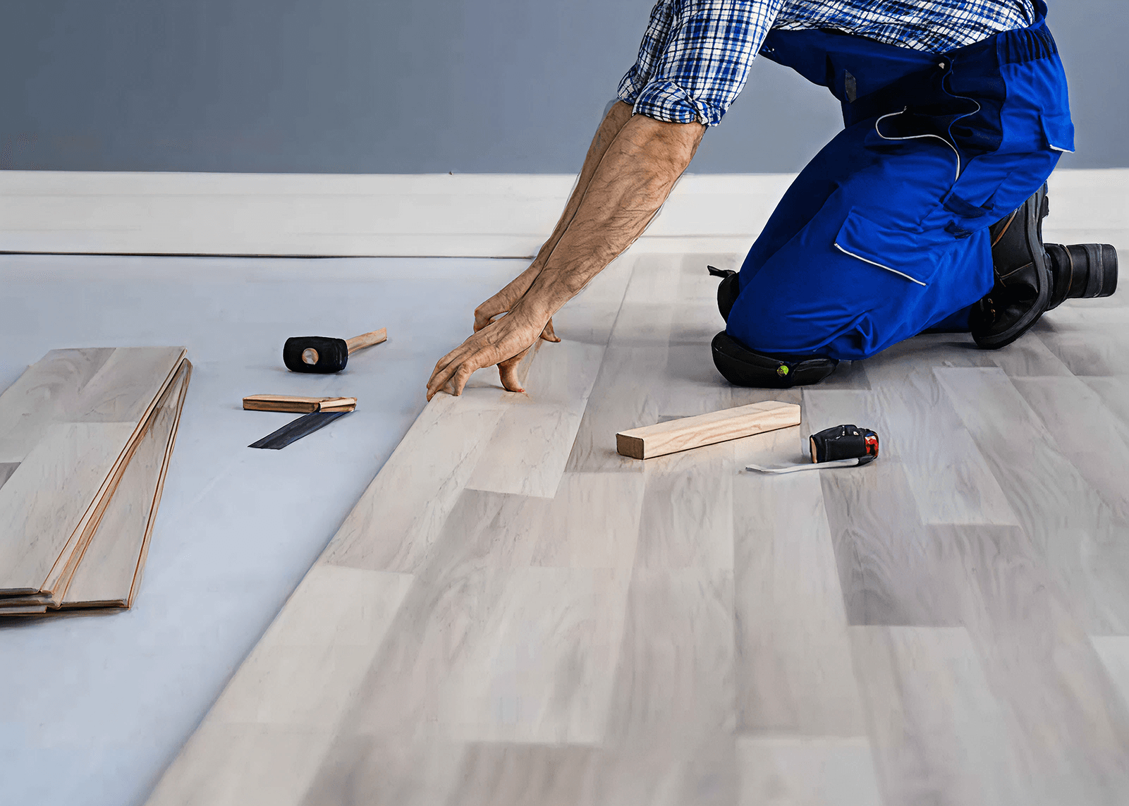 A person in blue overalls kneels on the floor, installing light wood laminate flooring. Tools, including a hammer and measuring tape, are scattered nearby.