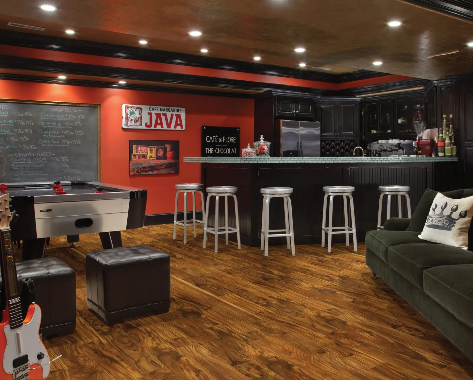Stylish basement with a cozy bar, four silver stools, retro signs, foosball table, dark cabinets, cushy green sofa, and warm wood flooring. Inviting ambiance.