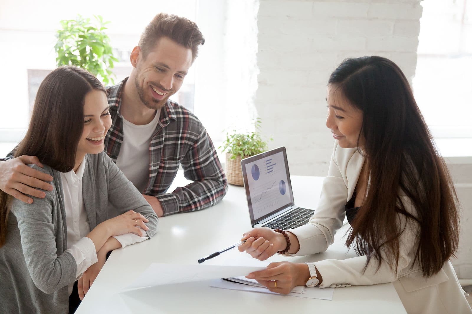 A couple smiles while consulting with a professional woman across a table. A laptop with graphs is open, creating a positive, collaborative atmosphere.