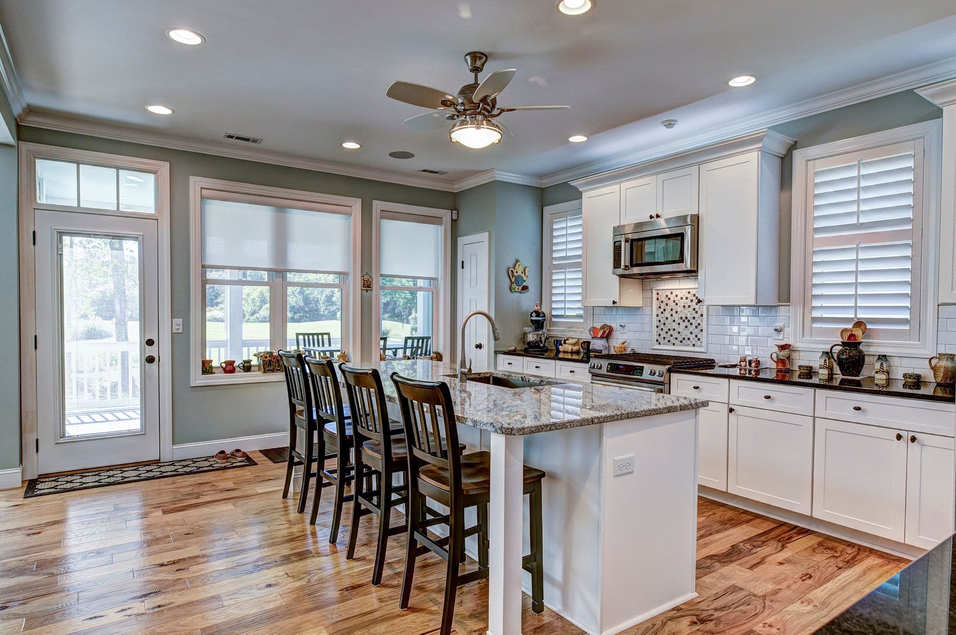 A spacious, modern kitchen with white cabinets, granite island seating four, and stainless steel appliances. Large windows and wood flooring add warmth.