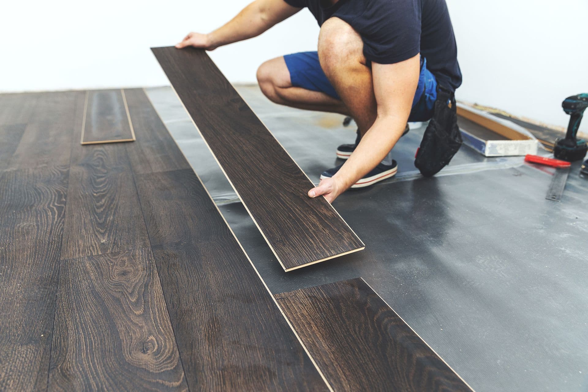 A person is installing dark wood flooring, placing a plank on a partially completed floor. Tools are visible, conveying a DIY home renovation scene.