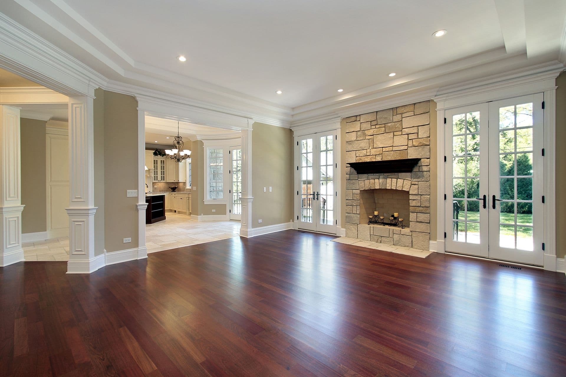 Bright, spacious living room with wooden floors, a large stone fireplace, and French doors opening to a garden. Adjacent kitchen visible.
