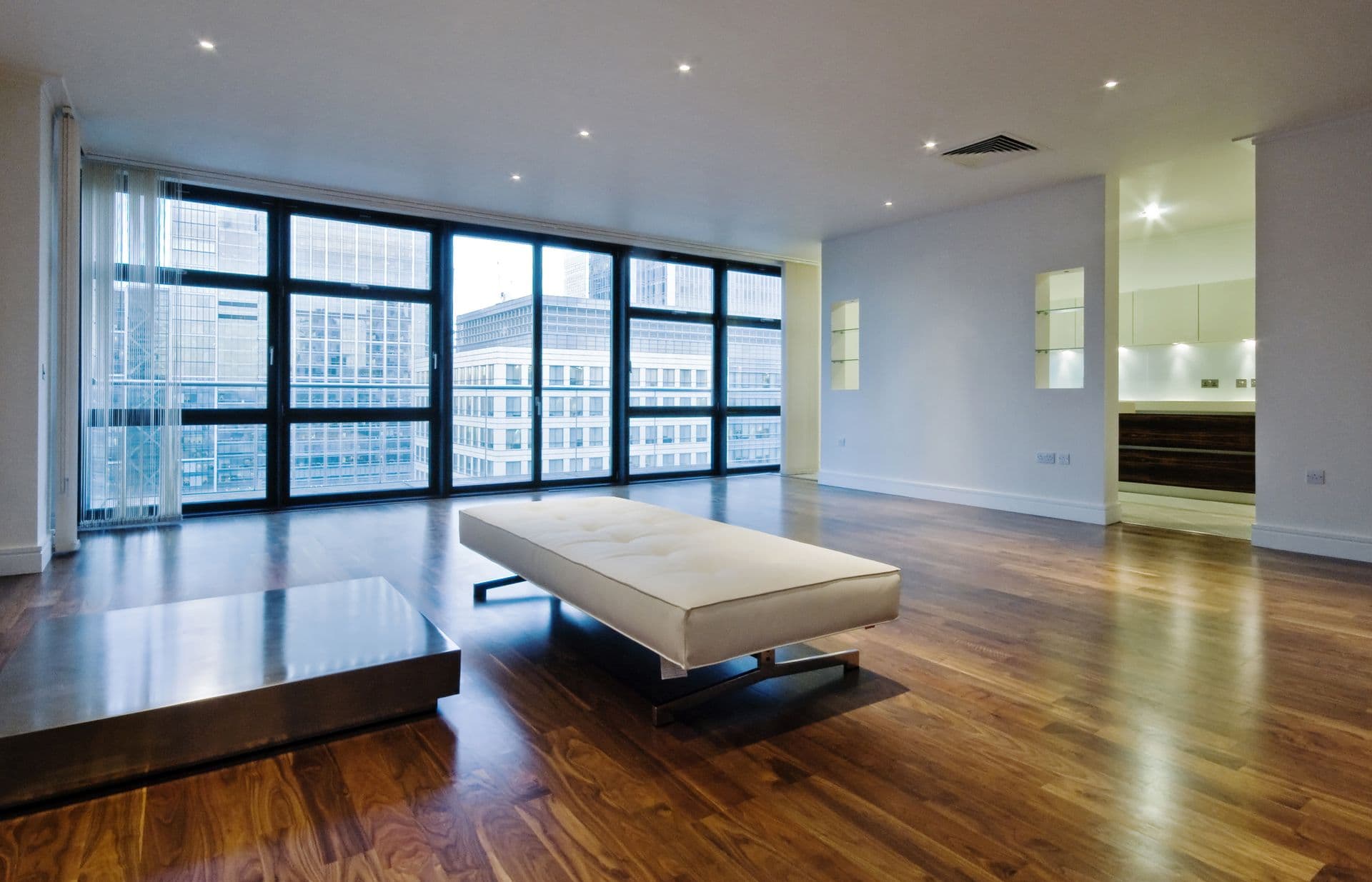 Spacious modern room with wooden floors, a white minimalist chaise lounge, ceiling lights, and large windows offering a cityscape view. Minimalist and serene.