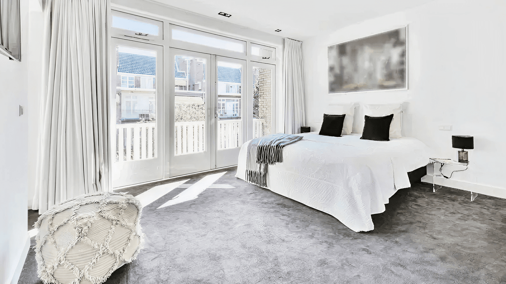 Bright, modern bedroom with white walls, a large bed with black pillows, a fluffy ottoman, and floor-to-ceiling windows letting in abundant light.
