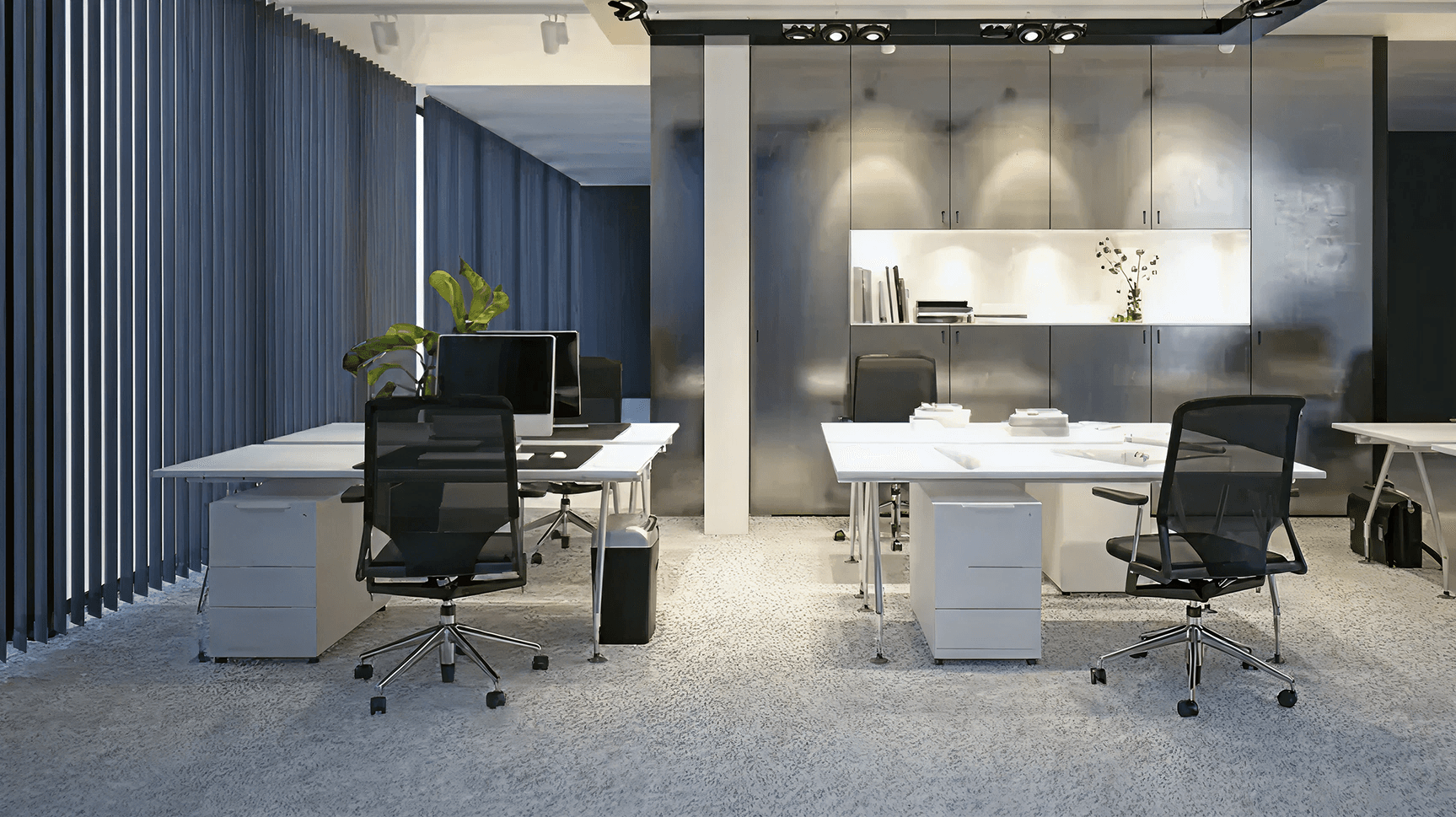 Modern office with sleek gray desks and chairs, indoor plants, and built-in cabinets. The space is well-lit and organized, conveying a professional tone.