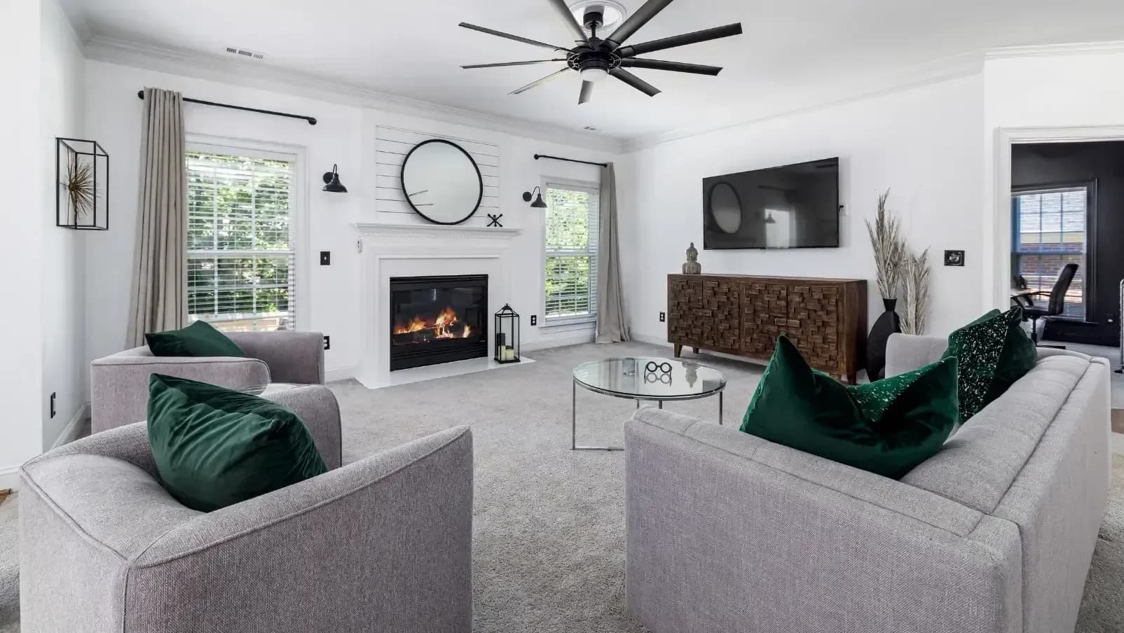 Modern living room with a cozy fireplace, round mirror above, and a large TV. Gray sofas with green pillows surround a glass coffee table. Peaceful ambiance.