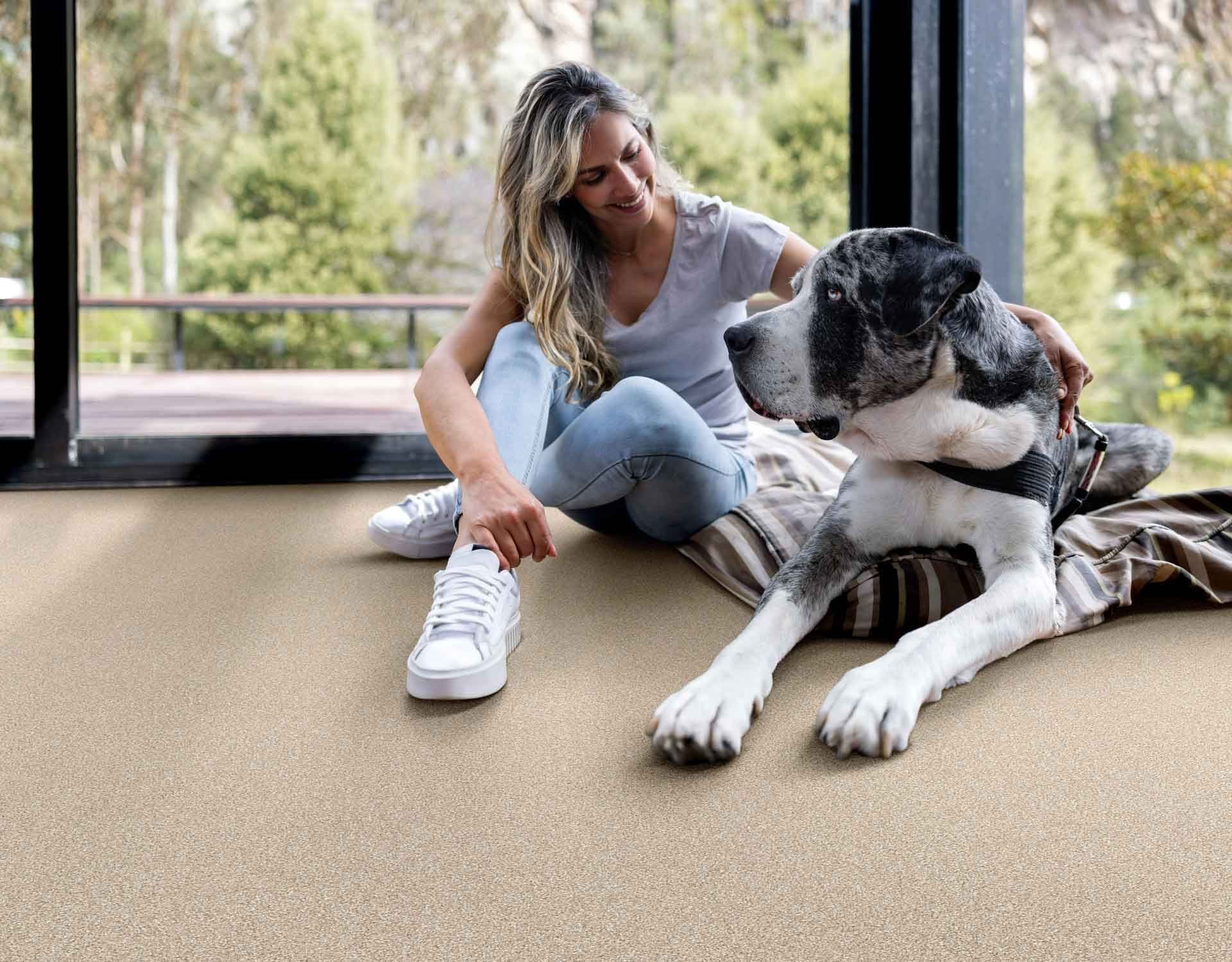 Woman sitting on a carpeted floor, smiling and petting a large grey and white dog lying beside her. Large windows show a lush garden outside.