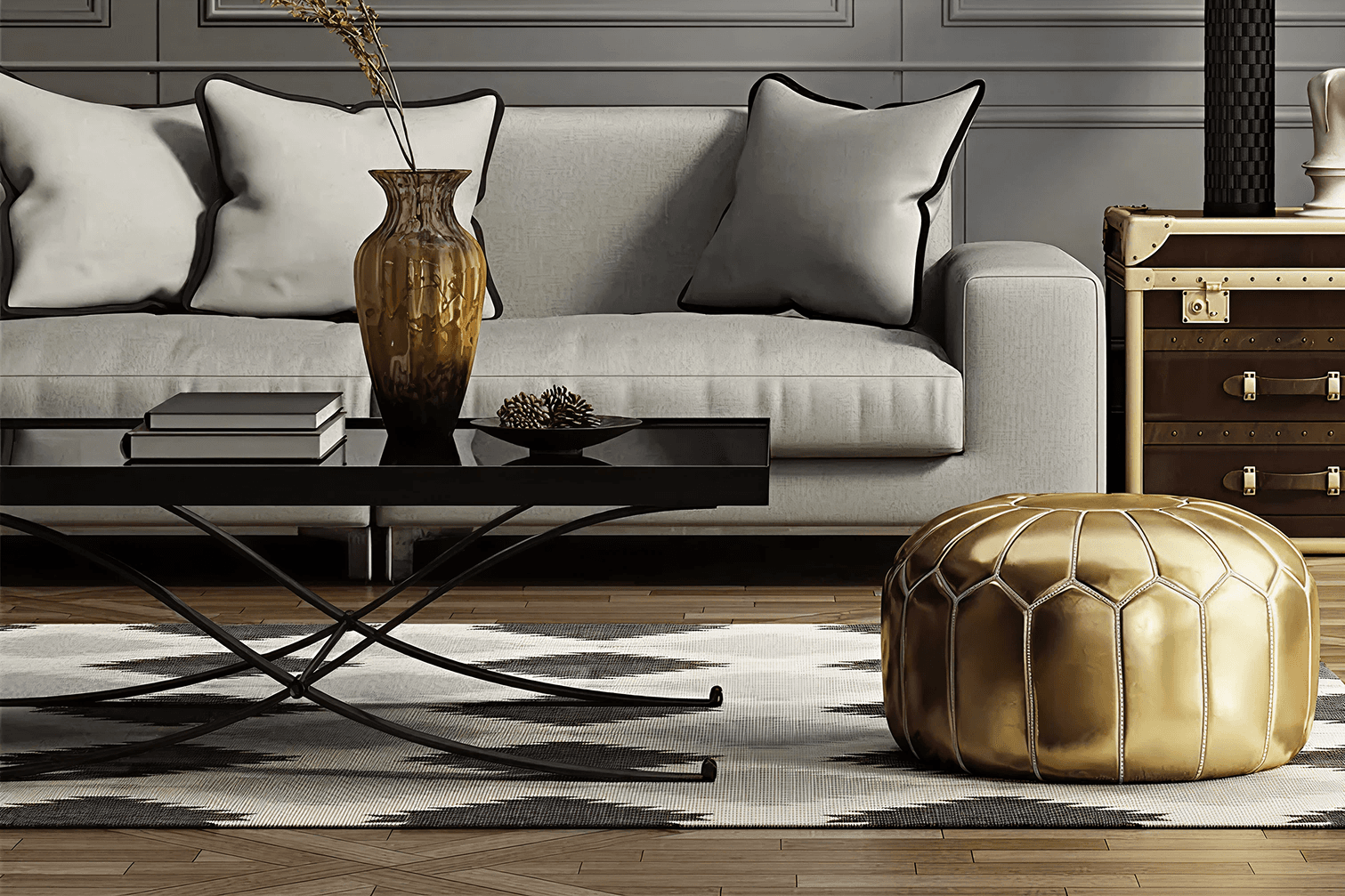 A modern living room with a gray sofa adorned with white pillows, a black coffee table holding books and a vase, and a gold pouf on a patterned rug.