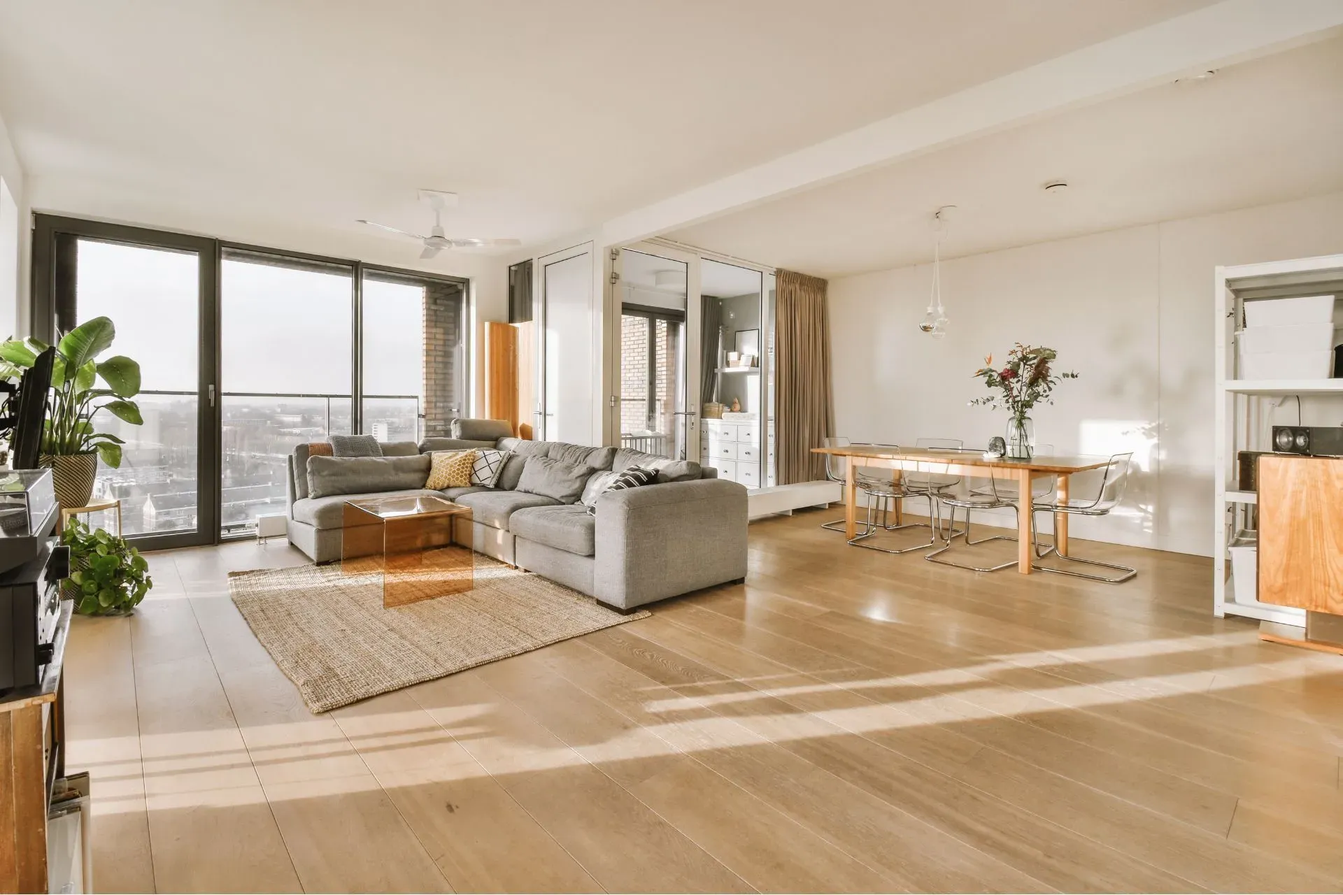 Spacious modern living room with large windows, abundant natural light, a gray sectional sofa, dining table, indoor plants, and wooden floors.
