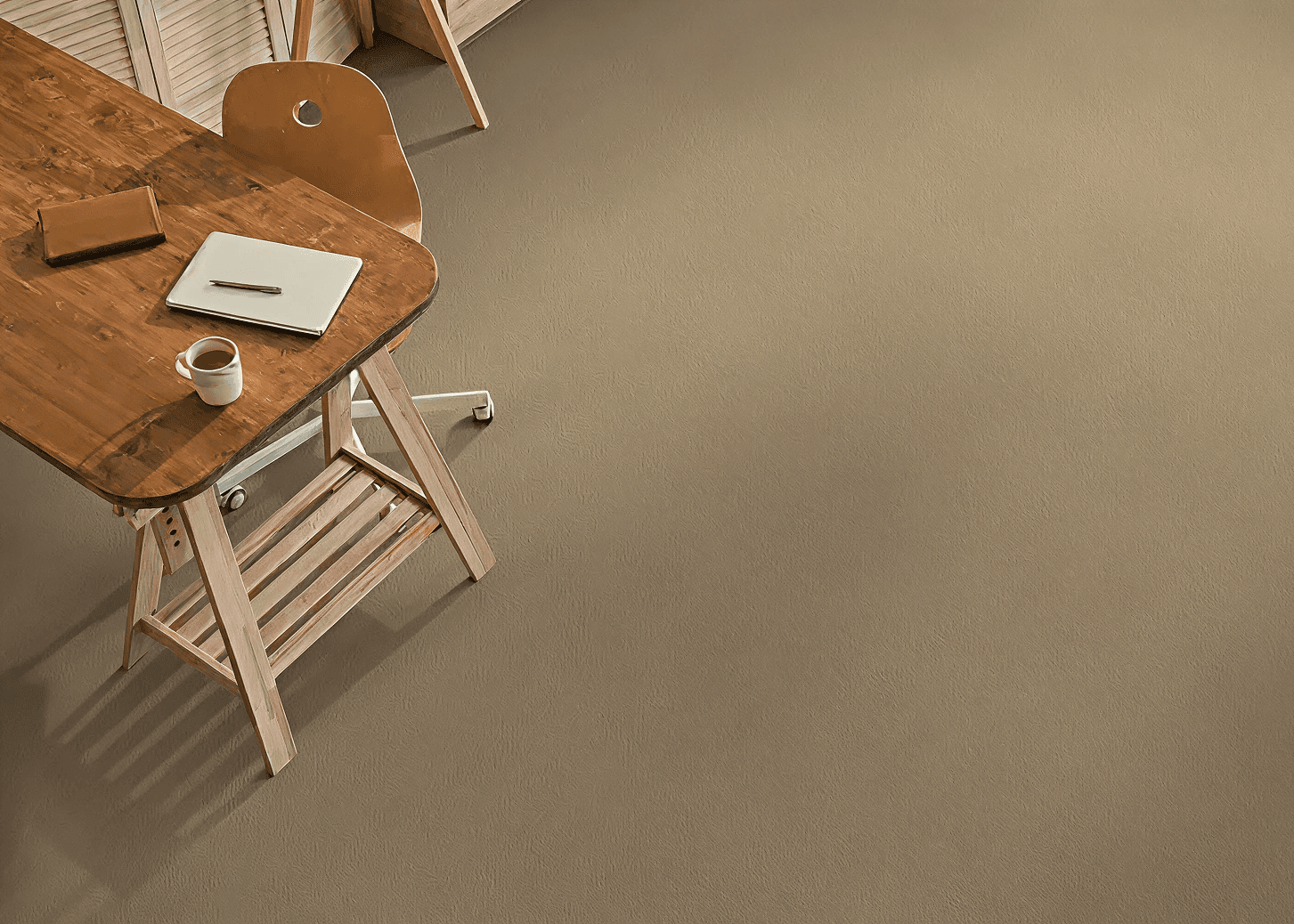 Wooden desk and chair on a beige carpet. Desk holds a notebook, closed laptop, pen, and coffee cup. Minimal, calm workspace conveys focus.