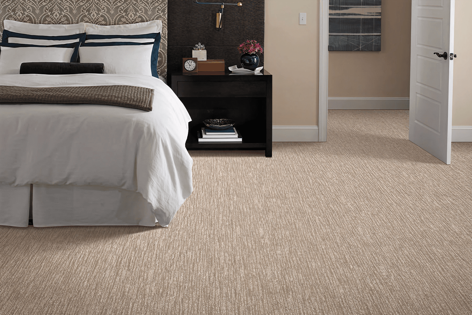 A cozy bedroom with a white bed, three pillows, and a dark blanket. A nightstand holds a clock and vase with flowers. An open door reveals a beige carpet.