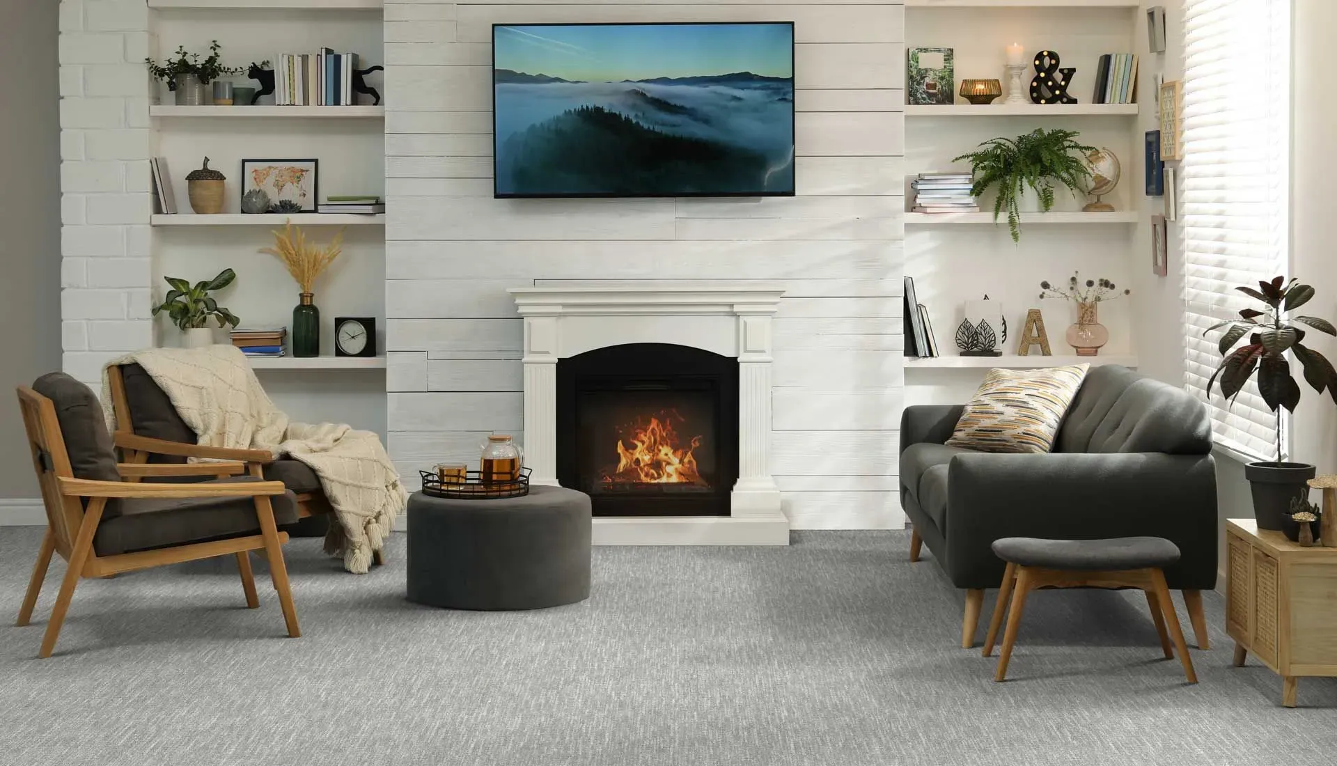 Cozy living room with a modern fireplace, gray armchairs, and a sofa. Shelves hold plants and decor, and a landscape painting hangs above the mantel.