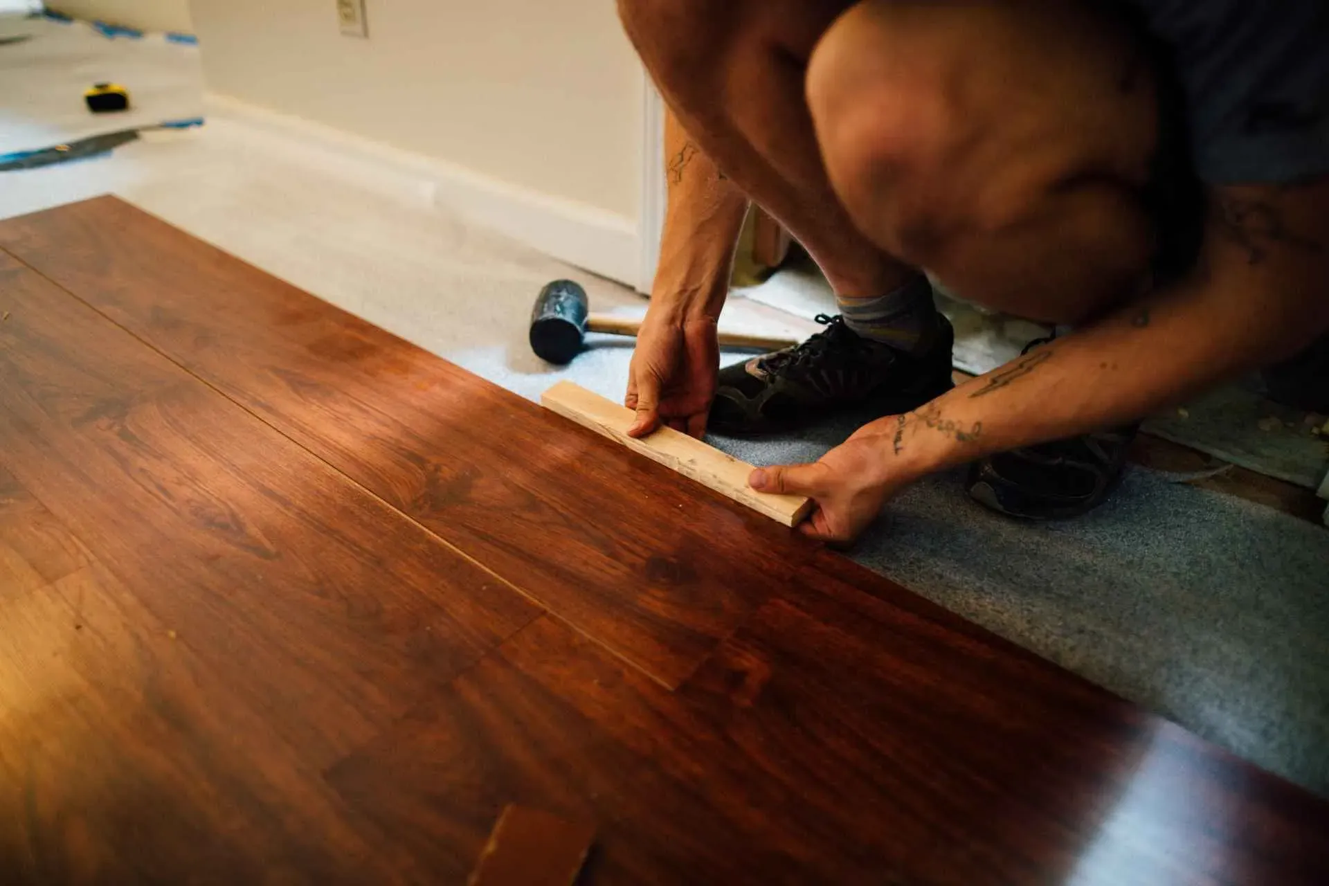 Person kneeling, installing hardwood flooring using a spacer, with a rubber mallet nearby on carpet. The scene conveys focus and manual work.