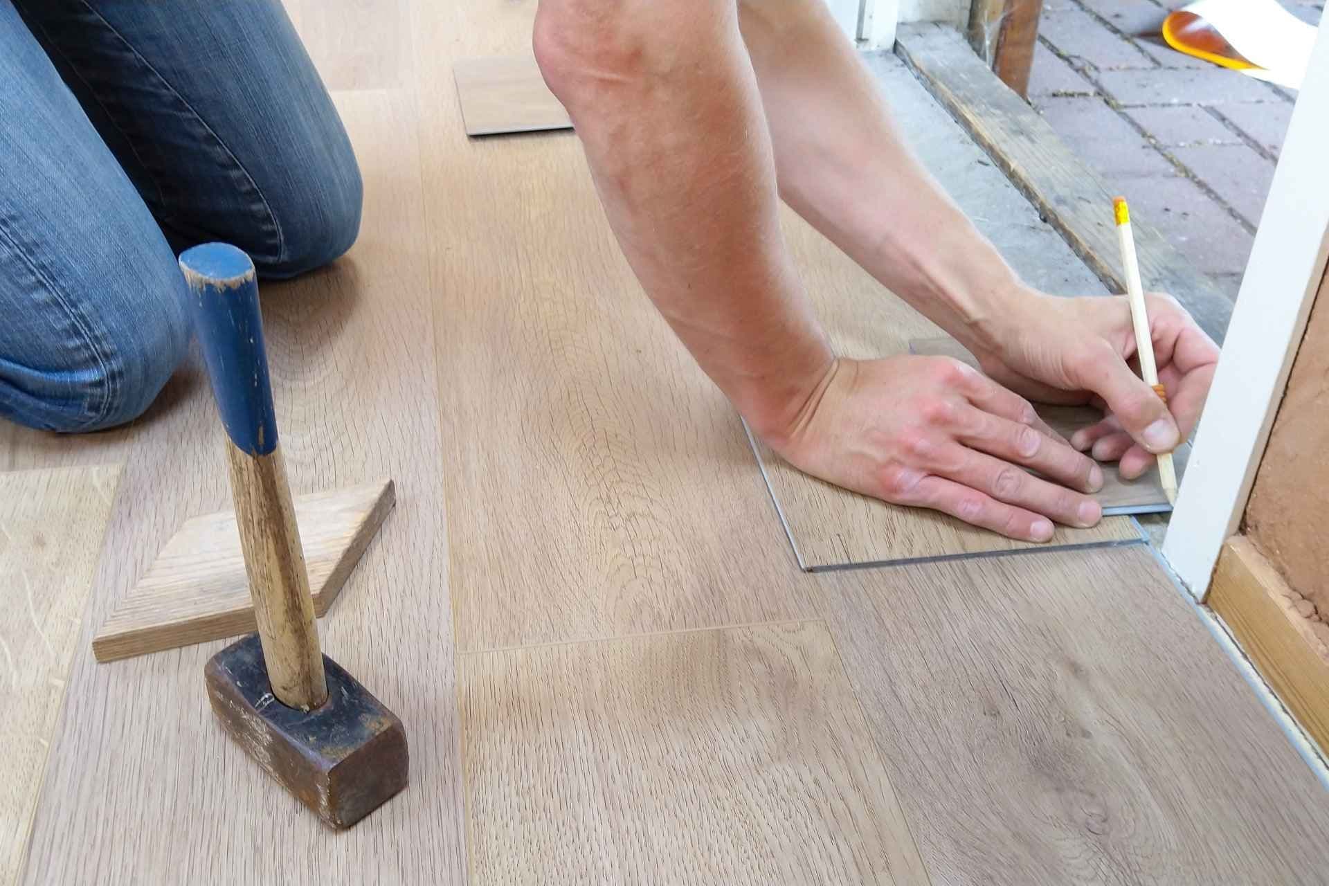 Person kneeling on light wood floor, measuring and marking a plank with a pencil. A wooden mallet rests nearby, suggesting a flooring project.