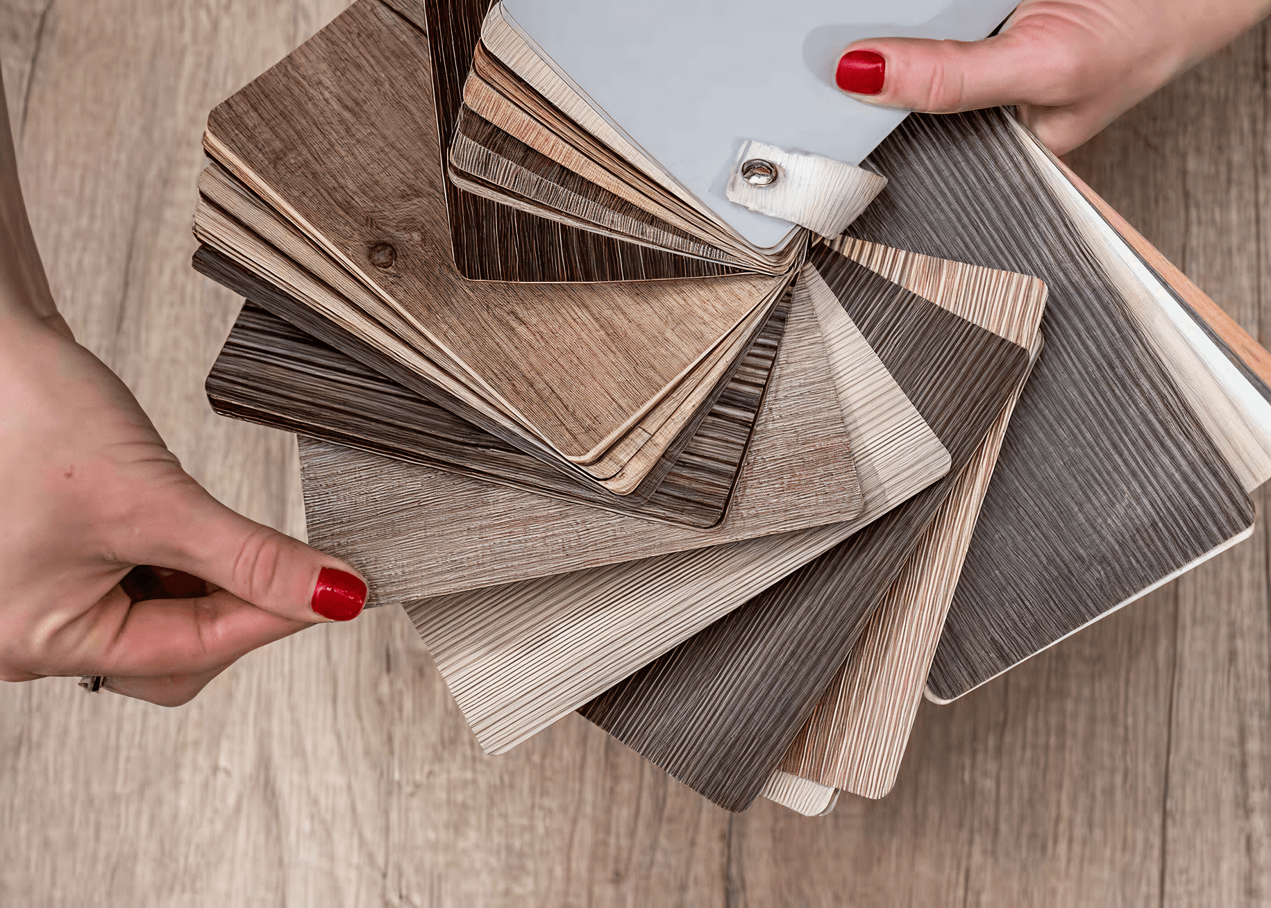 Hands displaying a fan of laminate flooring samples in various wood tones over a wooden floor, suggesting a choice or selection process.