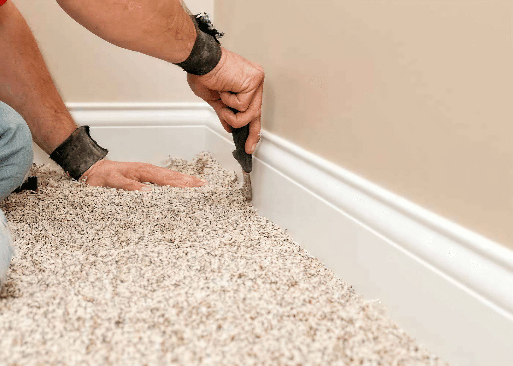 A person uses a carpet tucking tool against a white baseboard to fit a speckled carpet. The scene conveys focus and precision in home improvement.