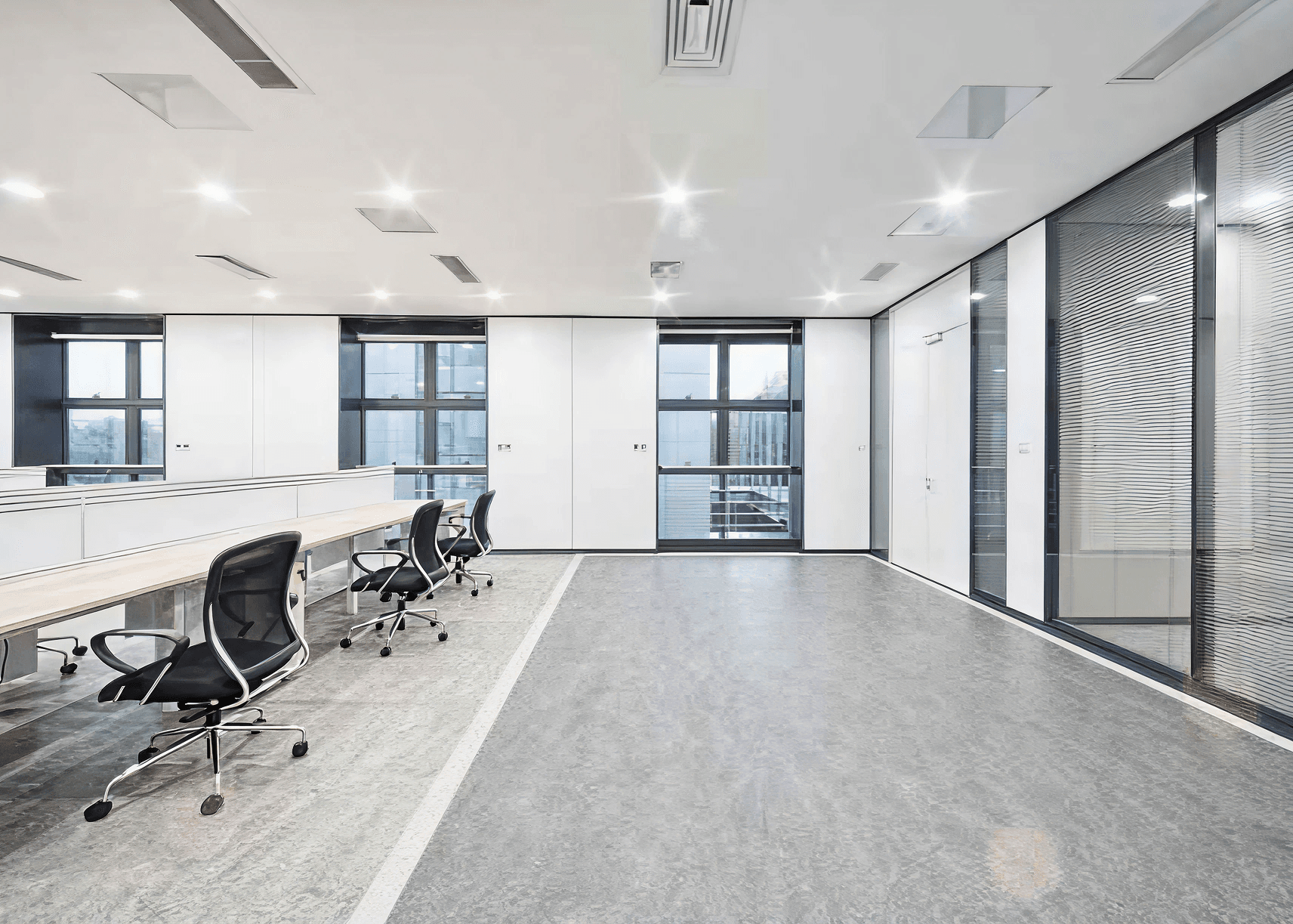 Modern office space with white walls and large windows. Empty desks with black chairs arranged in a row. Bright lighting creates a clean, minimalistic look.