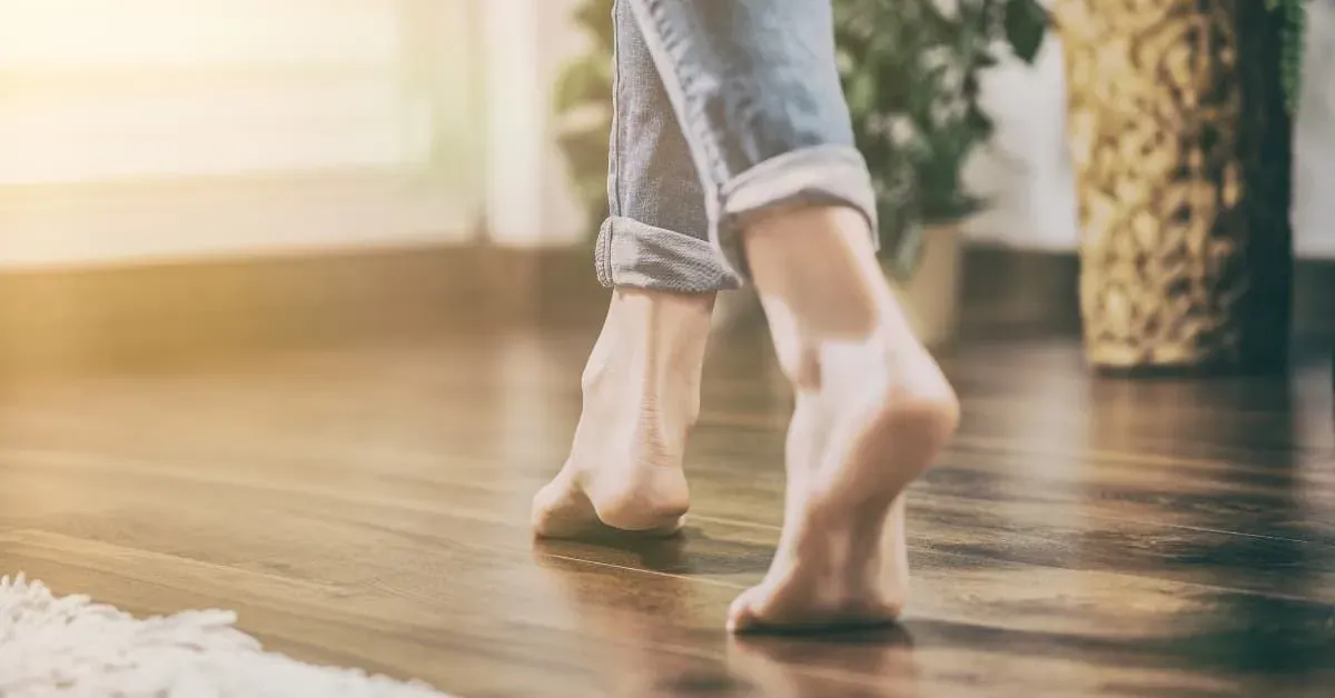 Bare feet walking on a sunlit wooden floor, with rolled-up jeans visible. Soft sunlight creates a warm, peaceful atmosphere. Plants and rug in the background.