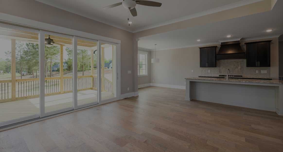 Spacious open-concept room with wooden floors, a ceiling fan, and sliding doors leading to a deck. Modern kitchen with dark cabinets in the background.