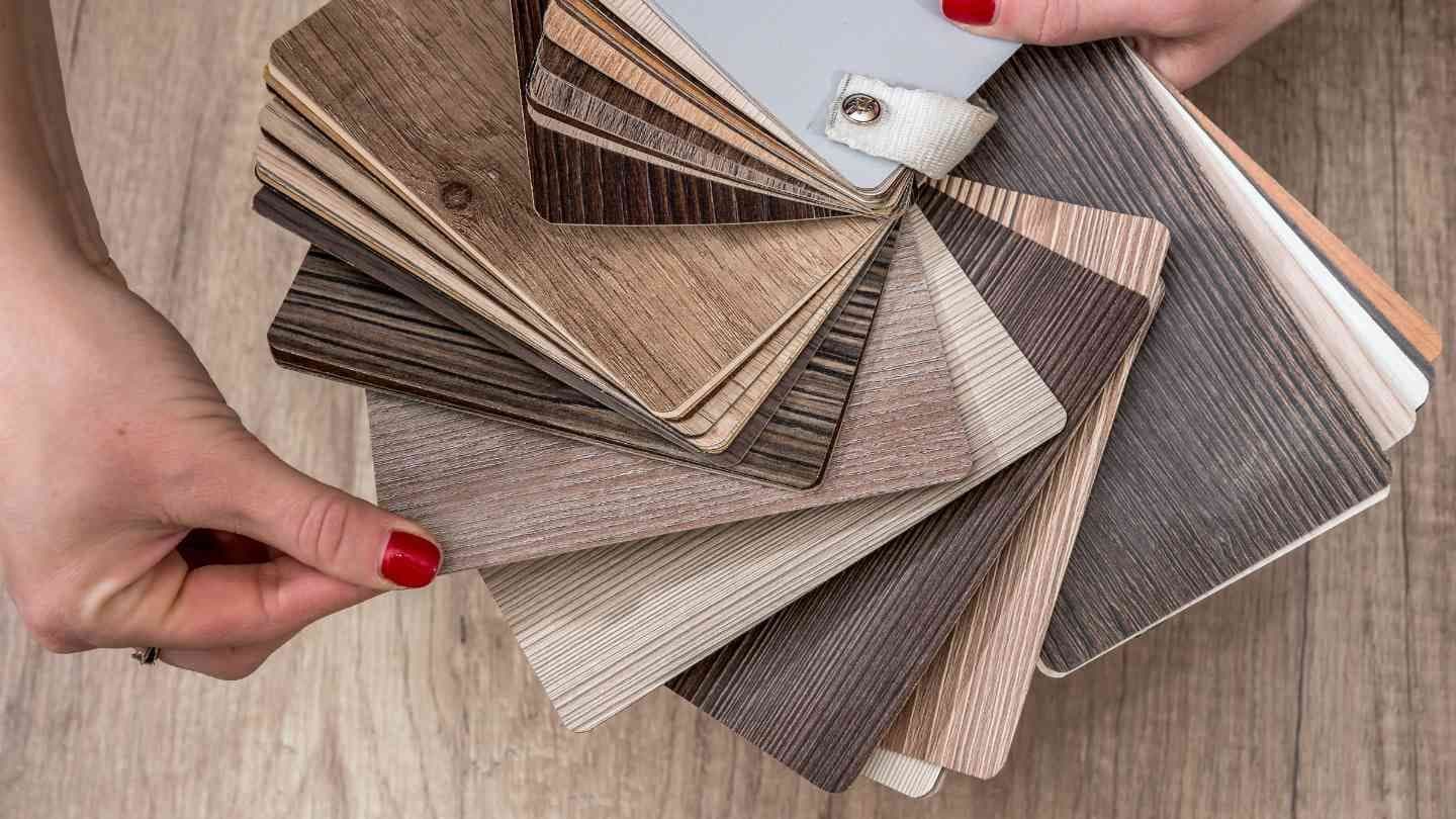 Hands with red nail polish hold a fan of wooden laminate samples in various shades and textures on a wooden surface, conveying choice and design.
