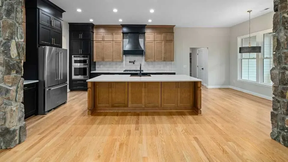 Spacious kitchen with wooden floors, a large island, and stone accents. Dark and light wood cabinets contrast with stainless steel appliances. Bright and modern.