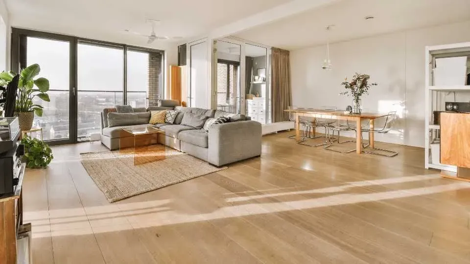 Bright, modern living room with wooden floors, gray sectional sofa, glass doors leading to a balcony, dining table with chairs, and green plants.