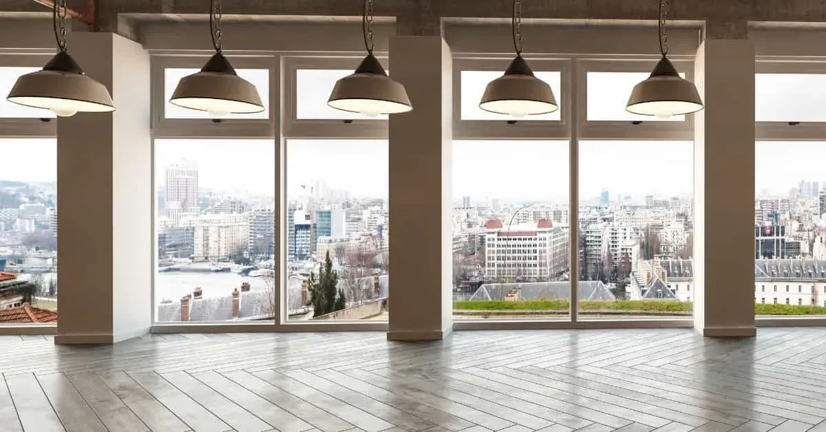 Spacious room with large windows overlooking a cityscape. Grey tiled floor, industrial pendant lights. Bright, open, and modern ambiance.