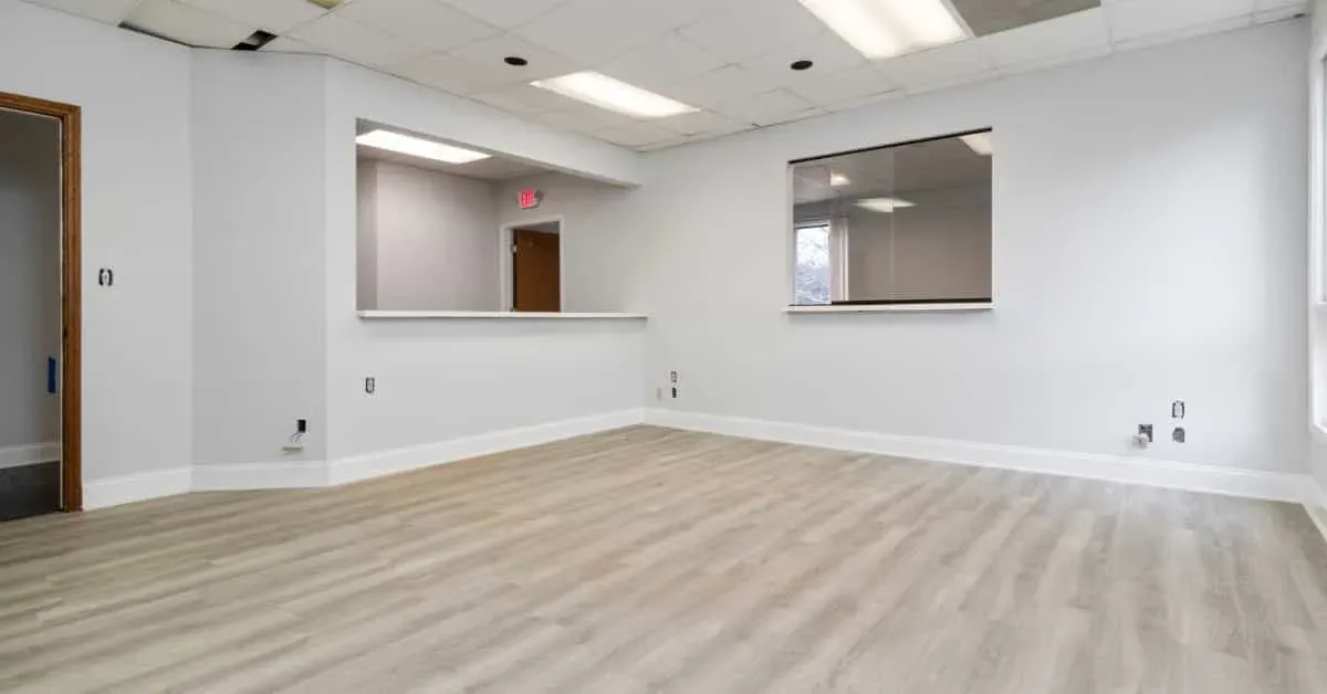 Spacious, empty office with light gray walls, wood flooring, and a drop ceiling. Features include a windowed reception area and a closed door. Bright lighting creates a clean atmosphere.