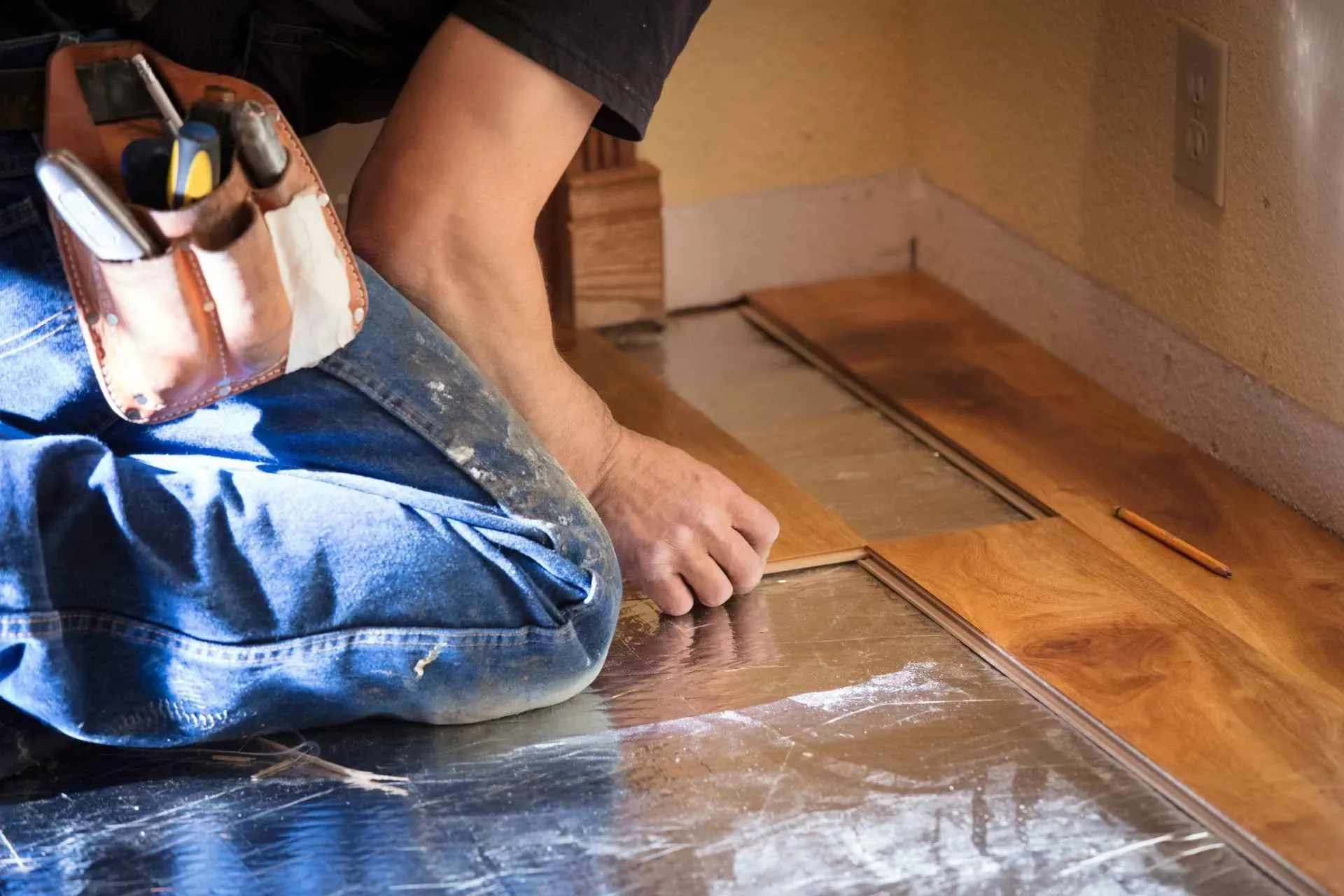 Person kneeling, installing wooden floor panels over reflective underlayment. Wearing blue jeans, tool belt with tools visible. Focused and precise atmosphere.