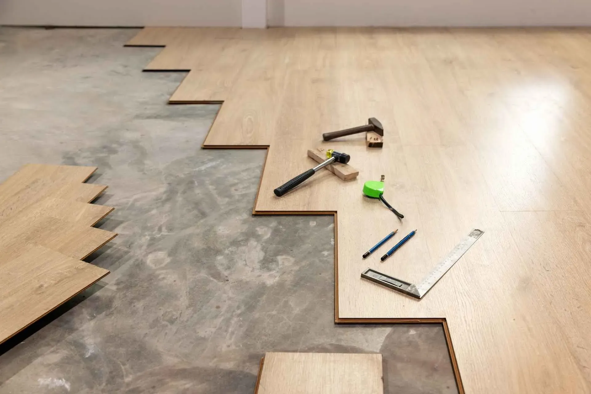 Wooden flooring installation in progress over concrete, showing tools like a hammer, tape measure, and pencil on laid planks, conveying a work-in-progress atmosphere.