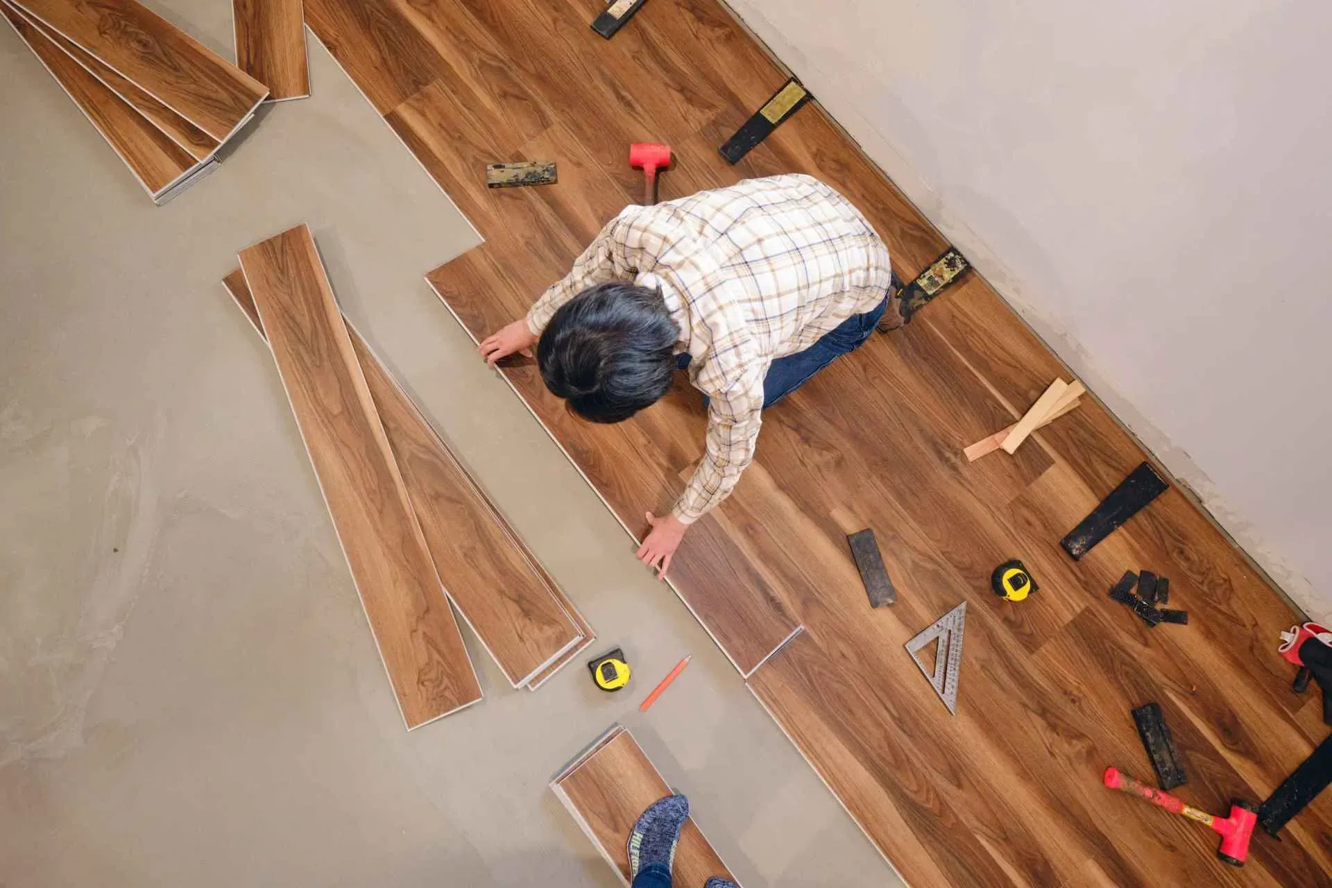 A person in a plaid shirt is installing wood laminate flooring in a room. Tools like a tape measure, square, and mallet are scattered around.