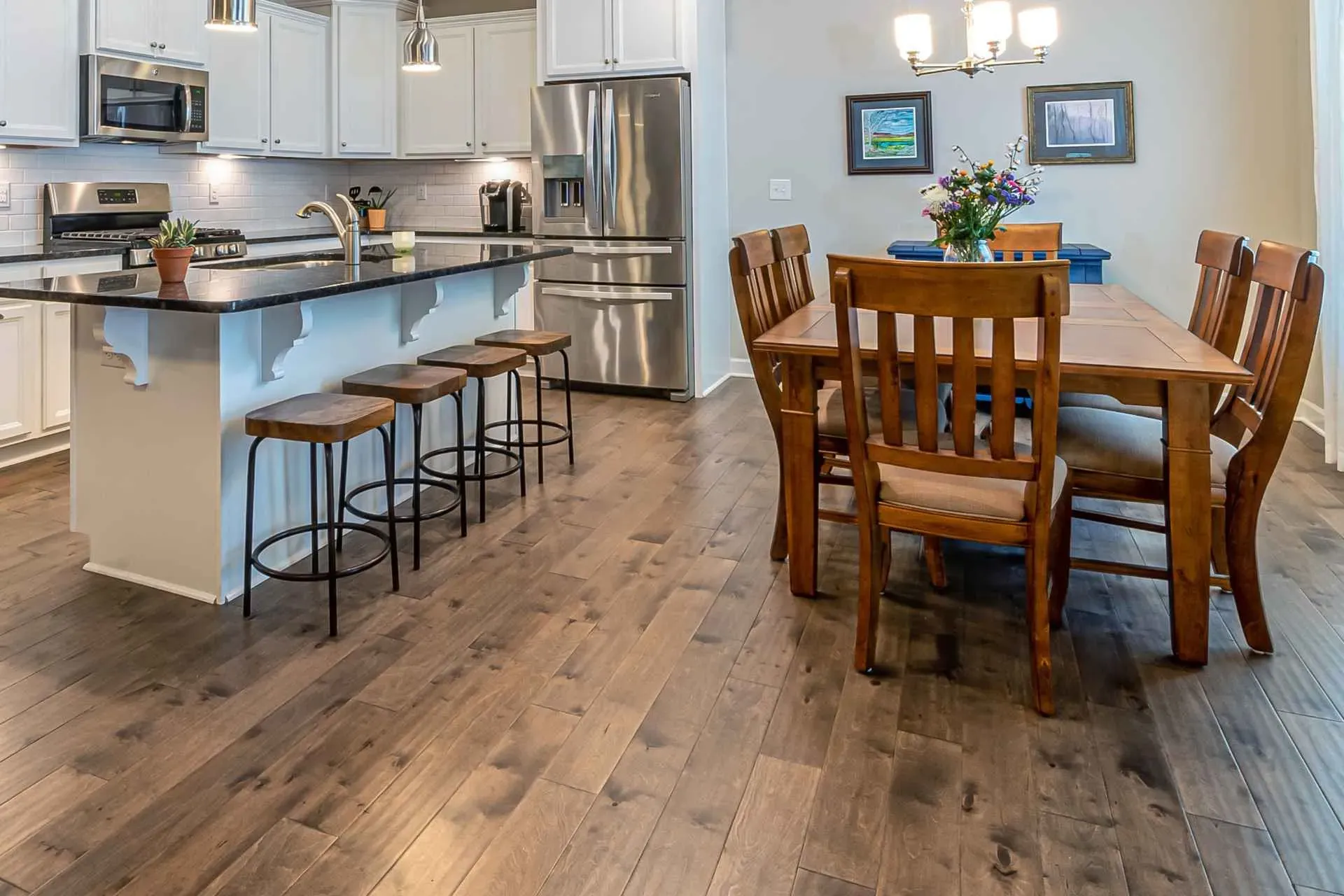 A modern kitchen and dining area with dark wood floors, featuring a granite island with stools, stainless steel appliances, and a wooden dining table.