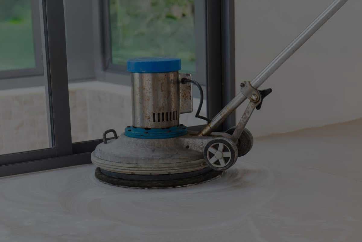 A floor polishing machine with a blue top is shown cleaning a smooth, light-colored surface near large windows. The scene feels tidy and industrious.