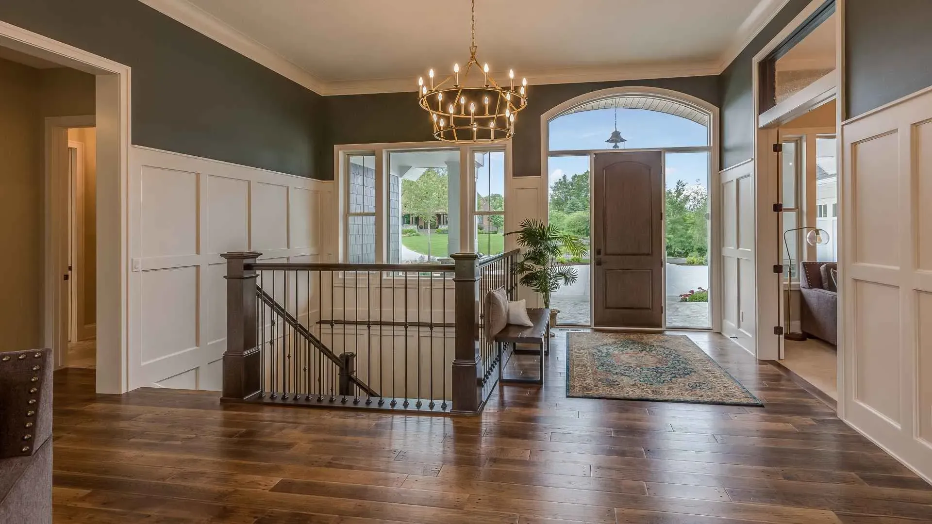 Elegant foyer with hardwood floors, a chandelier, and white wainscoting. A staircase leads down, and a large window shows a lush garden view.