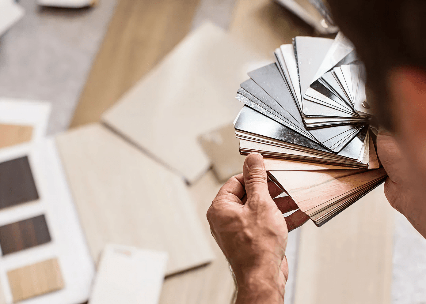 Hands holding a fan of laminate samples in wood and metallic finishes over a table scattered with more samples. The mood is focused and creative.