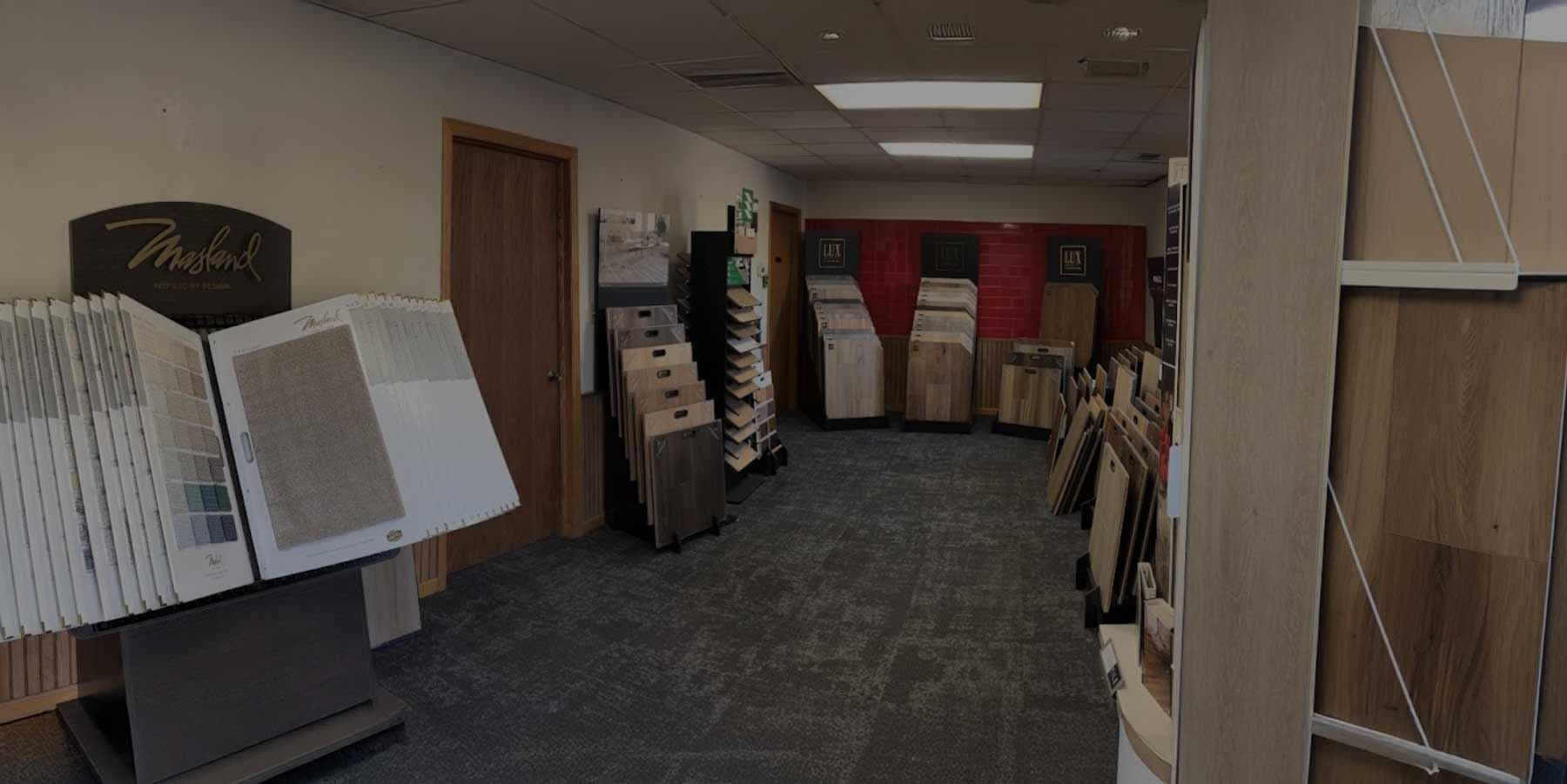 A showroom with various wood flooring samples arranged on displays against walls and in racks. The room has a carpeted floor and bright lighting.