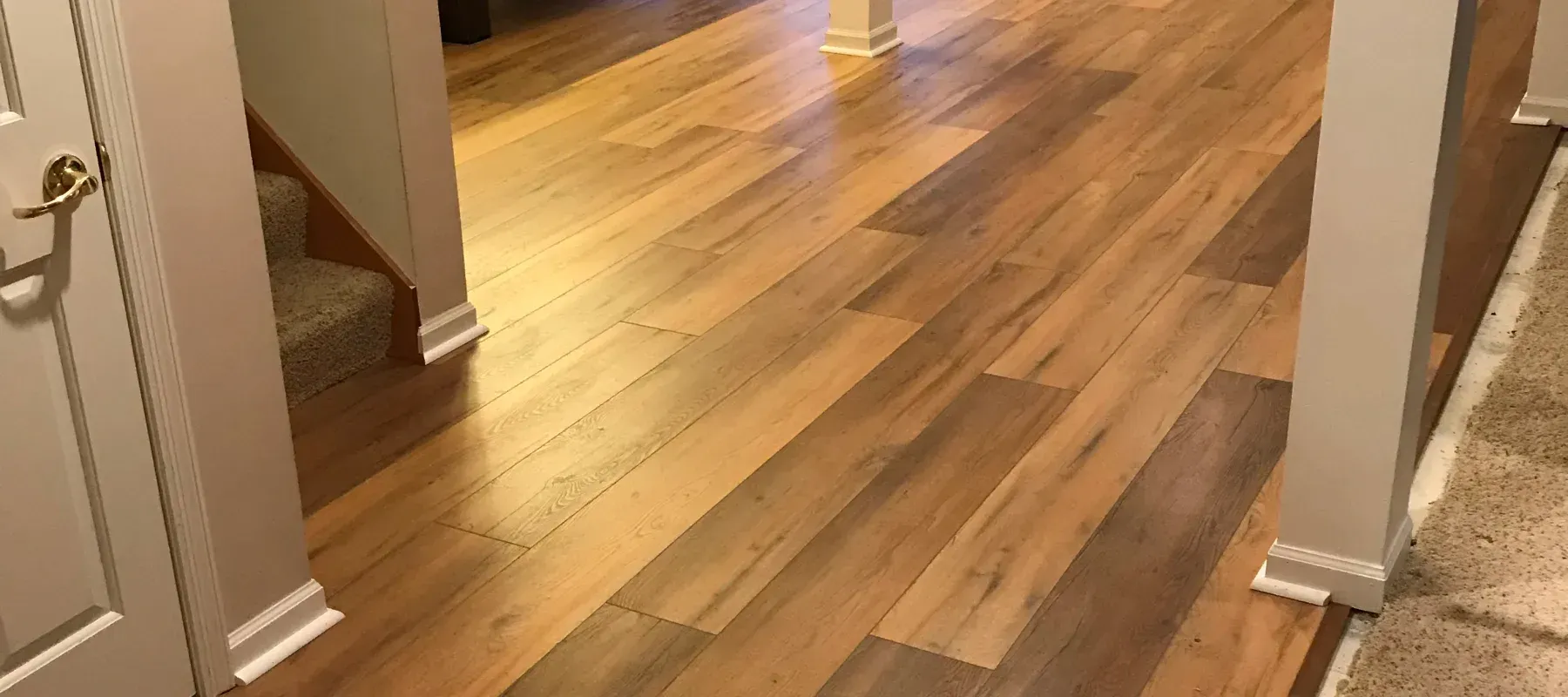 Alt text: Warm-toned laminate flooring extends through a well-lit hallway, flanked by white walls and columns. Stairs with carpeting are visible on the left.