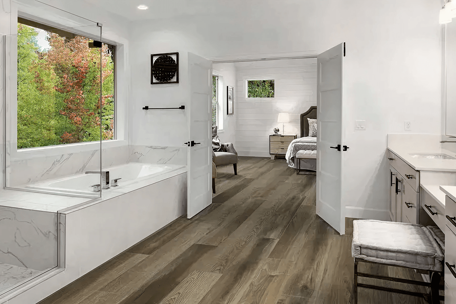 Spacious bathroom with white marble tub, large window showing greenery, and wooden floor leading to a cozy bedroom with open double doors.