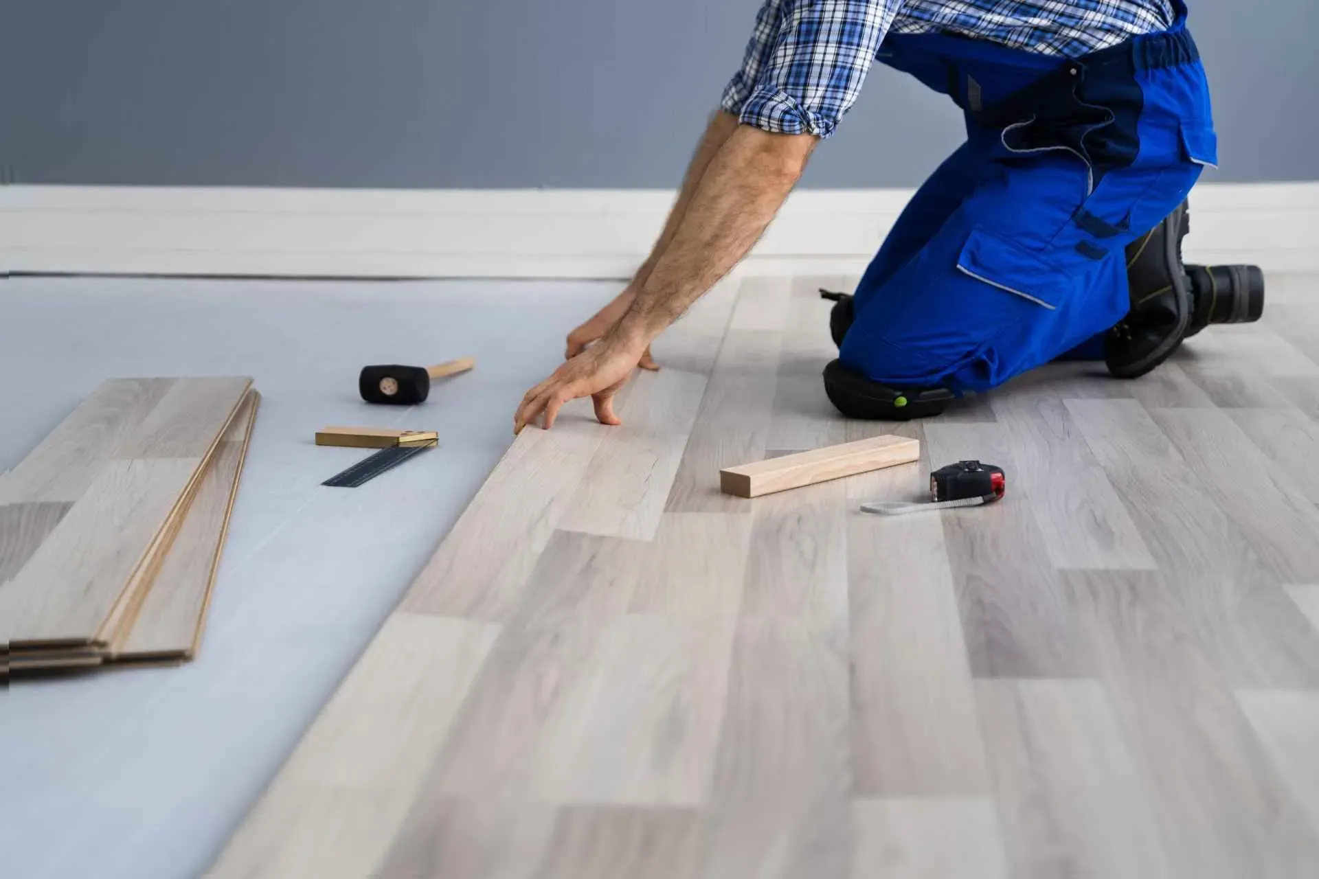 A person in blue overalls is installing light wood laminate flooring, using tools like a mallet and measuring tape on a gray background.