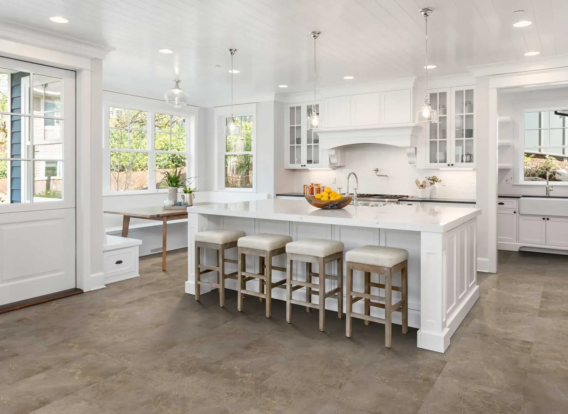 Bright kitchen with white cabinetry and marble countertops, featuring a large island with four stools. Pendant lights hang above, and a bowl of oranges adds color. Large windows let in natural light, enhancing a clean and inviting atmosphere.
