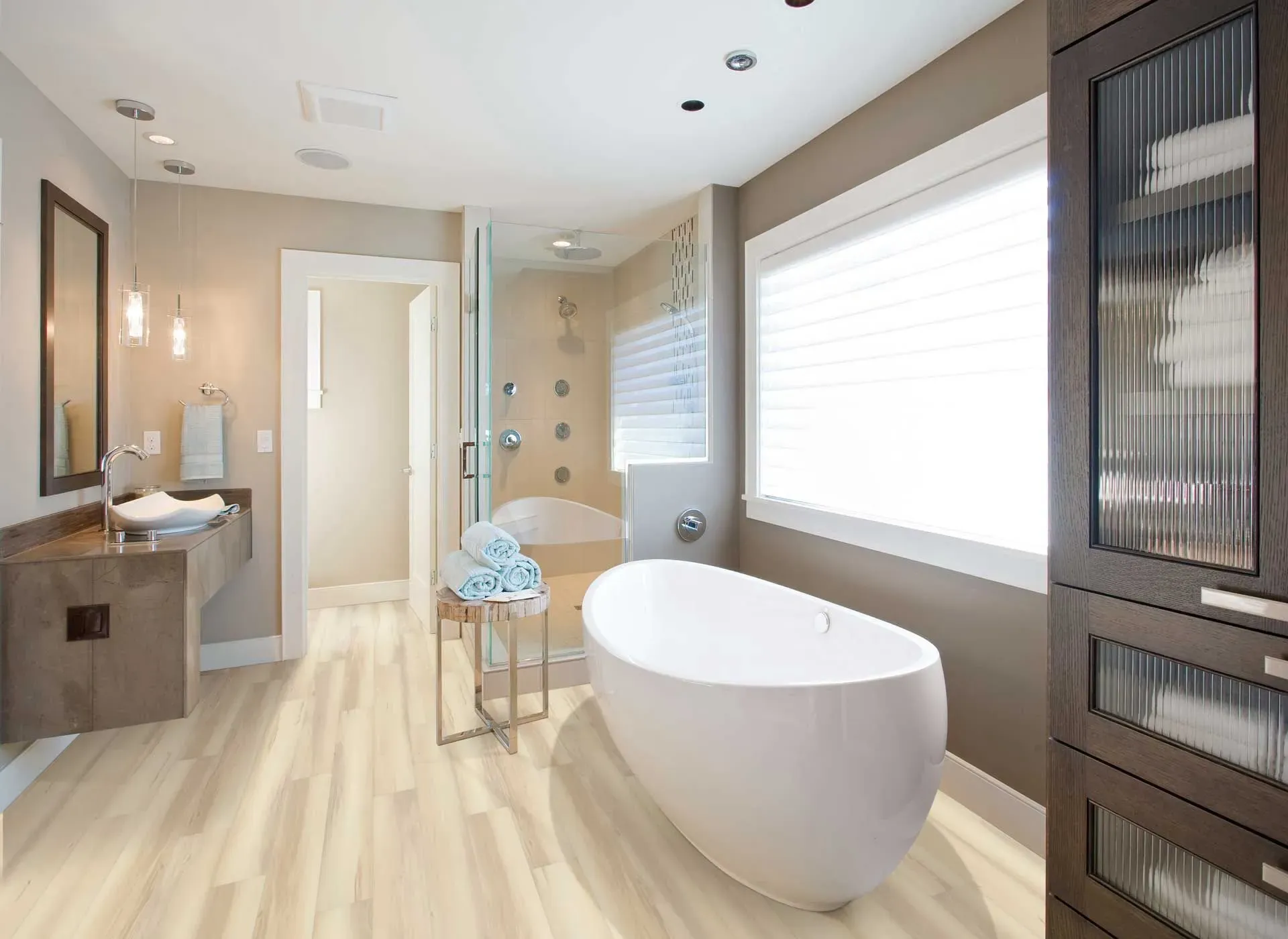 Modern bathroom with a freestanding tub, large window with blinds, glass-enclosed shower, floating vanity, pendant lights, and soft wood flooring.