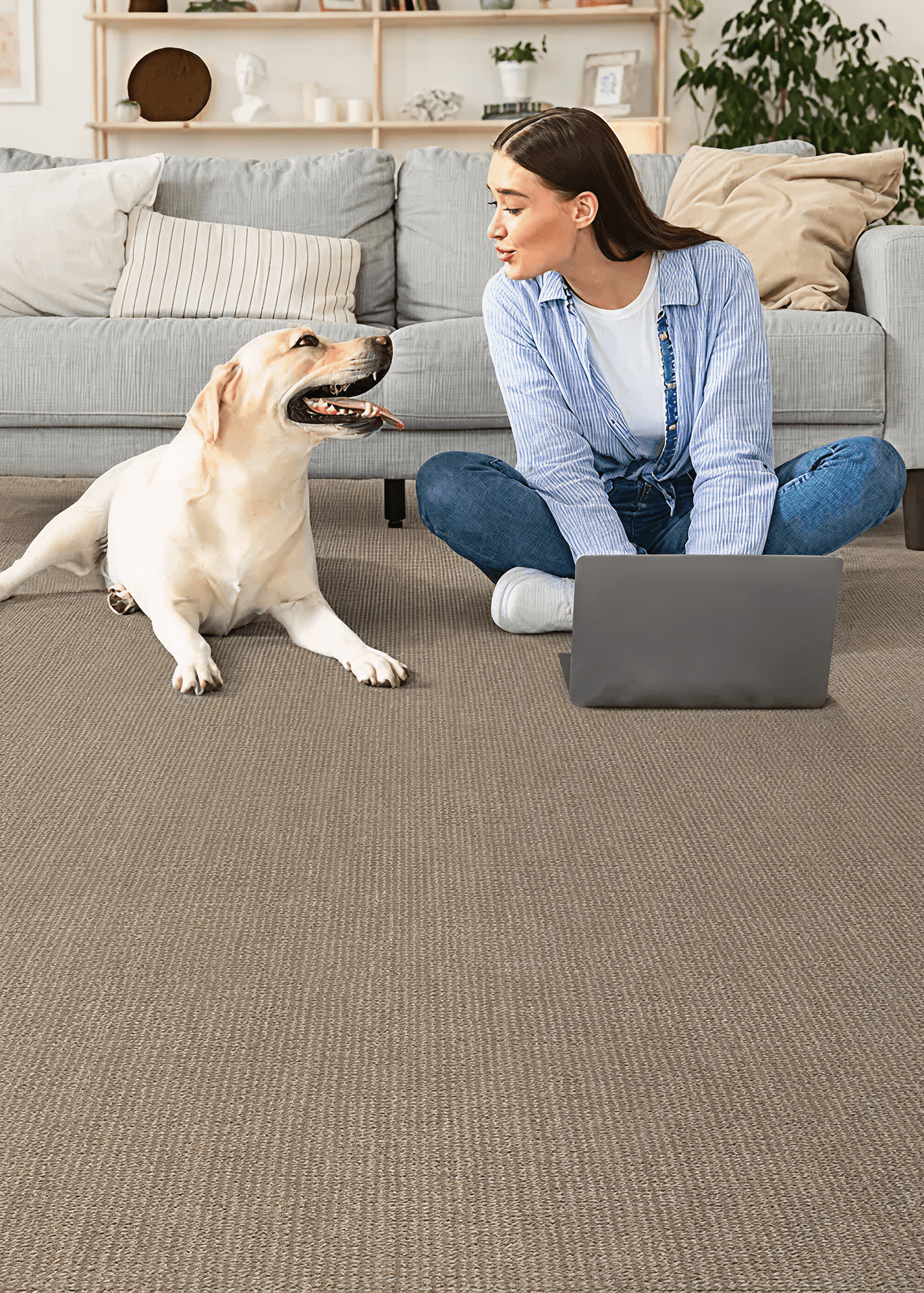 A woman sits on a carpeted floor with a laptop, smiling at a Labrador next to her. A cozy grey sofa and shelves with decor are in the background.