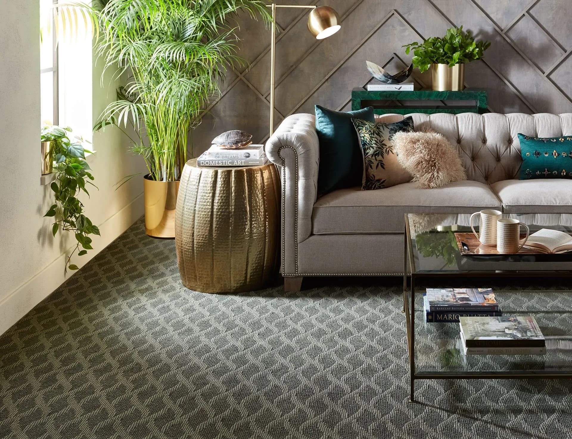 Stylish living room with a light gray tufted sofa, green pillows, gold side table, lush plants, geometric wall pattern, and glass coffee table.