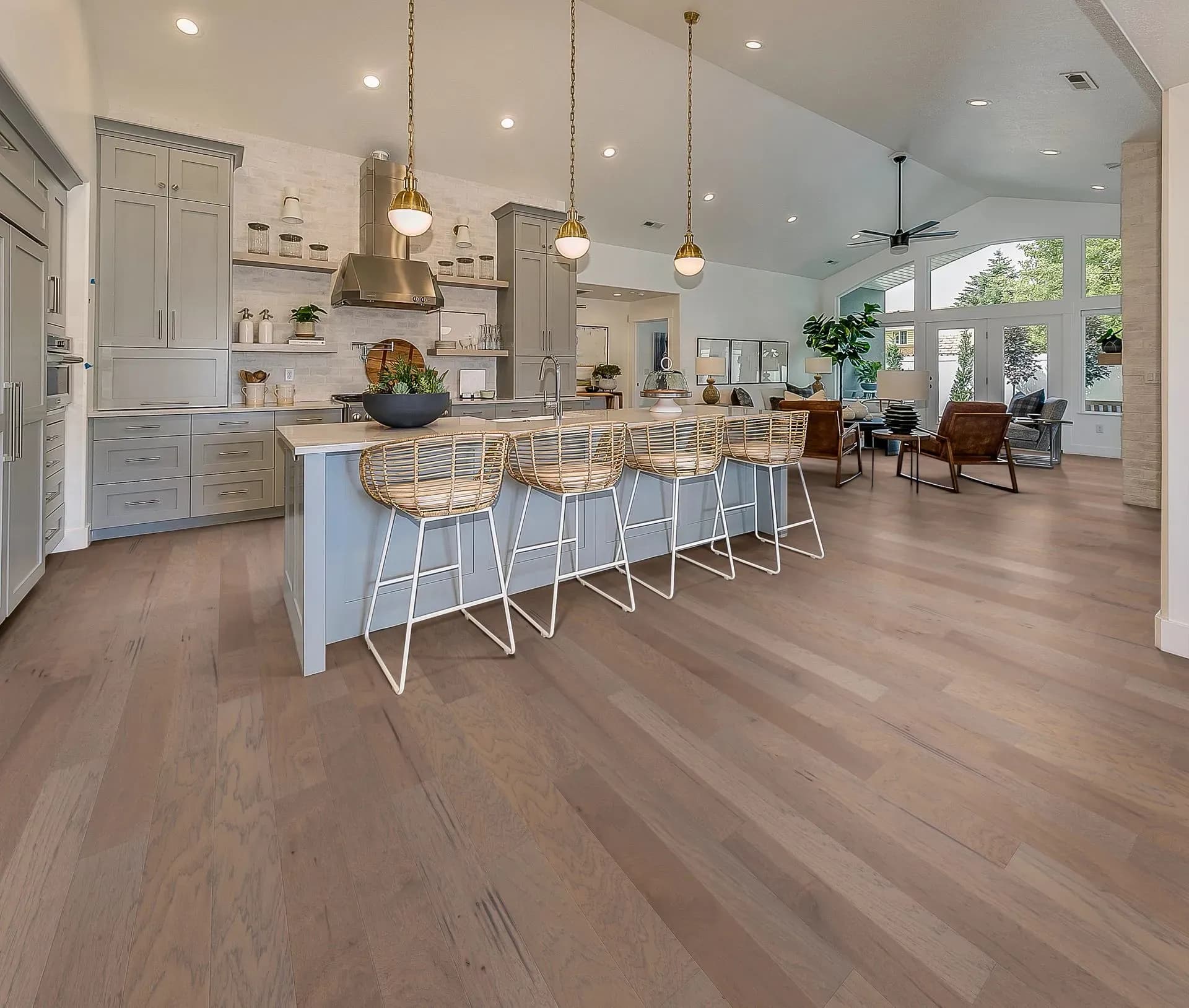 Spacious kitchen and living area with light wood floors, gray cabinets, a large island with rattan chairs, pendant lights, and modern decor. Airy and inviting.