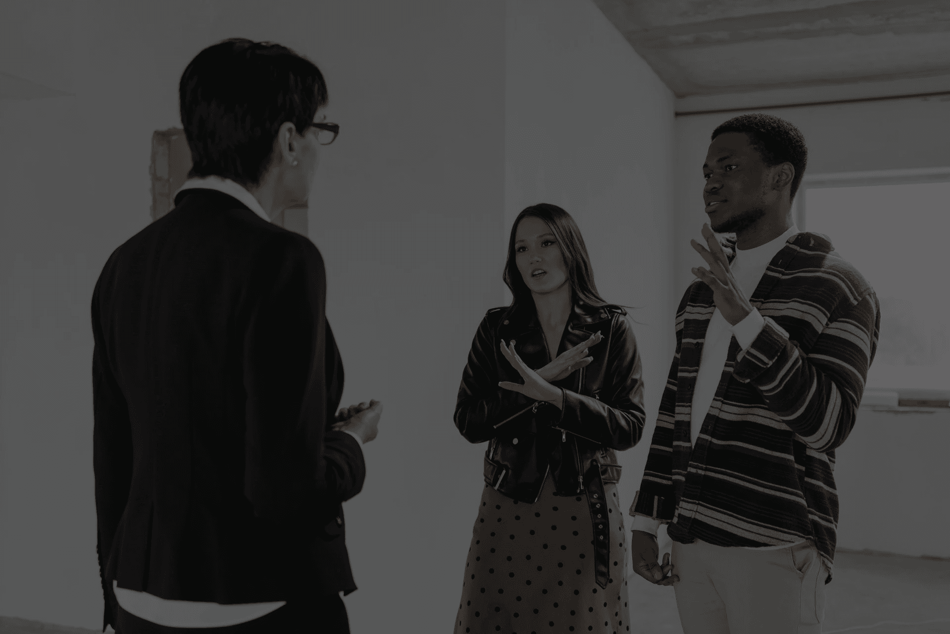 Three people stand in a white-walled room discussing something. Two are gesturing expressively, while one listens attentively. The mood is engaged.