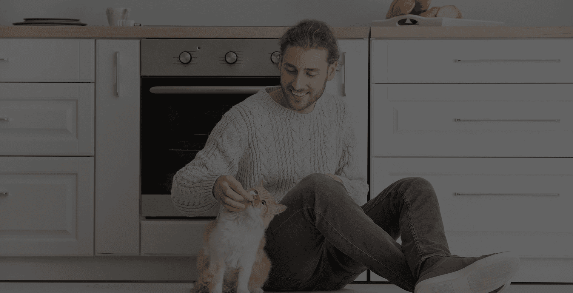 Man with long hair and a knitted sweater sits on the kitchen floor, smiling at an orange and white cat. The kitchen is modern with white cabinets.