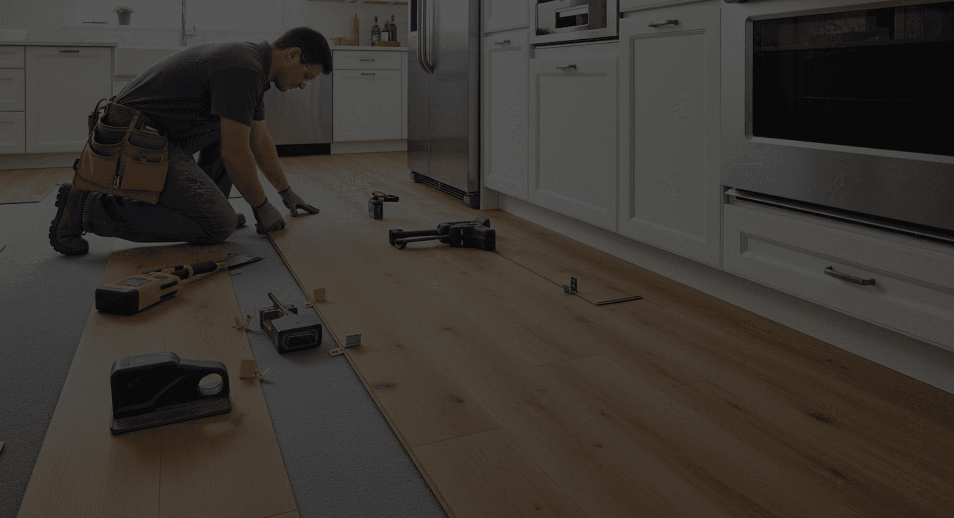 A man kneels in a modern kitchen installing light wood flooring. Tools are scattered on the floor, and white cabinets create a bright, clean atmosphere.