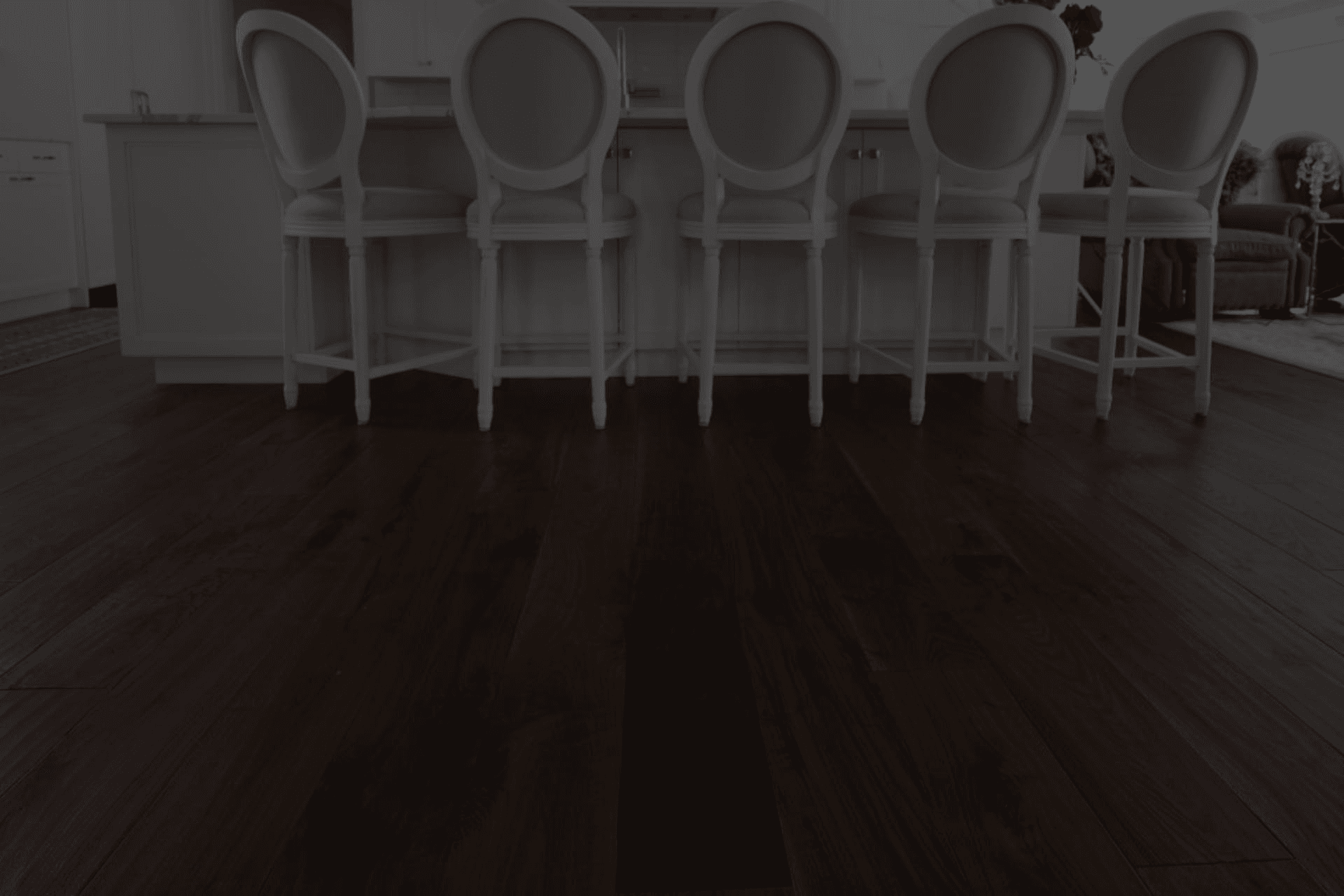 Elegant kitchen area with dark hardwood floors and classic white barstools facing a sleek counter. A cozy seating area with gray chairs is visible.