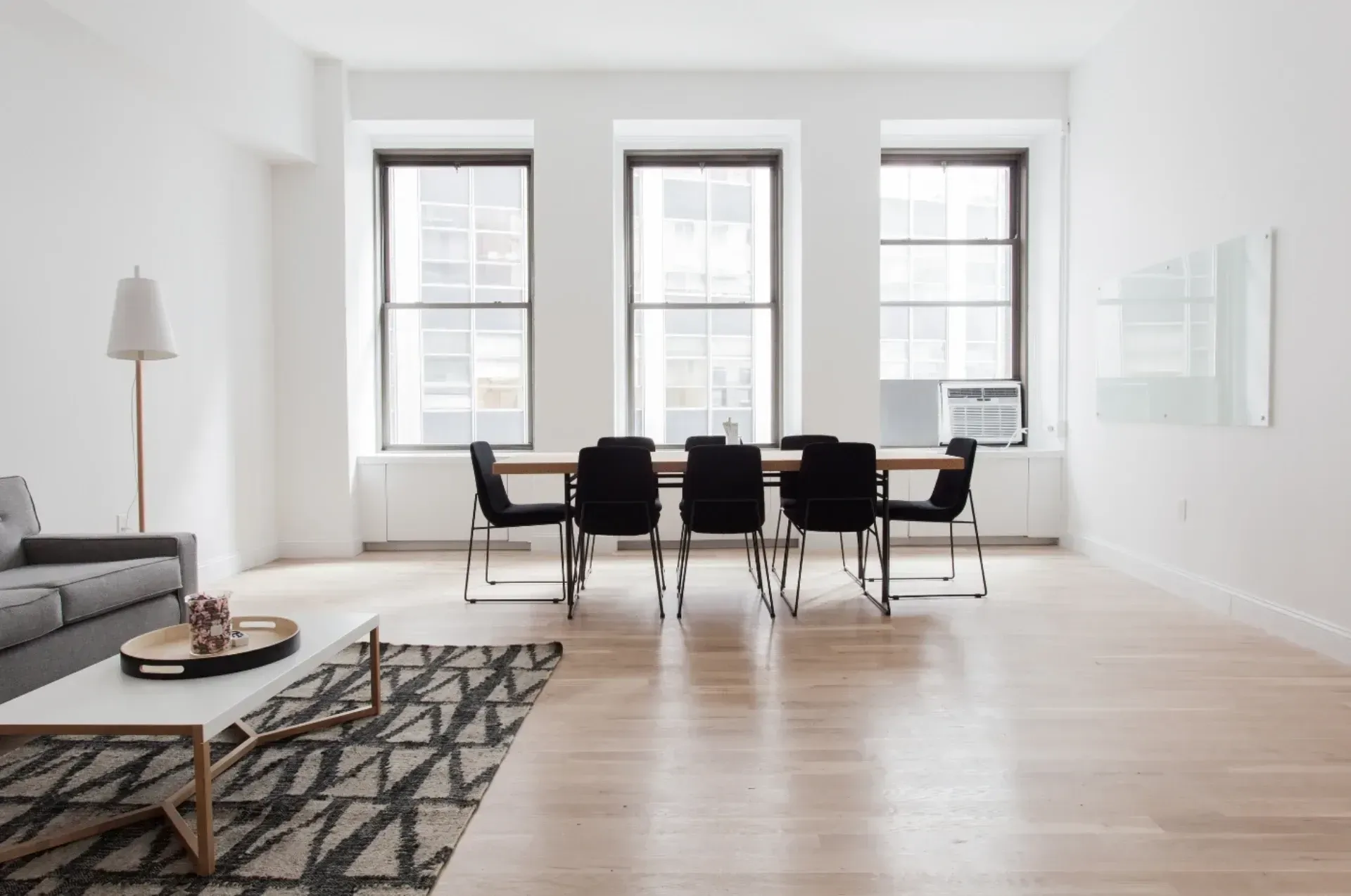 Minimalist room with large windows, a black dining table, and chairs. A gray sofa and geometric rug add style. Bright and airy ambiance.