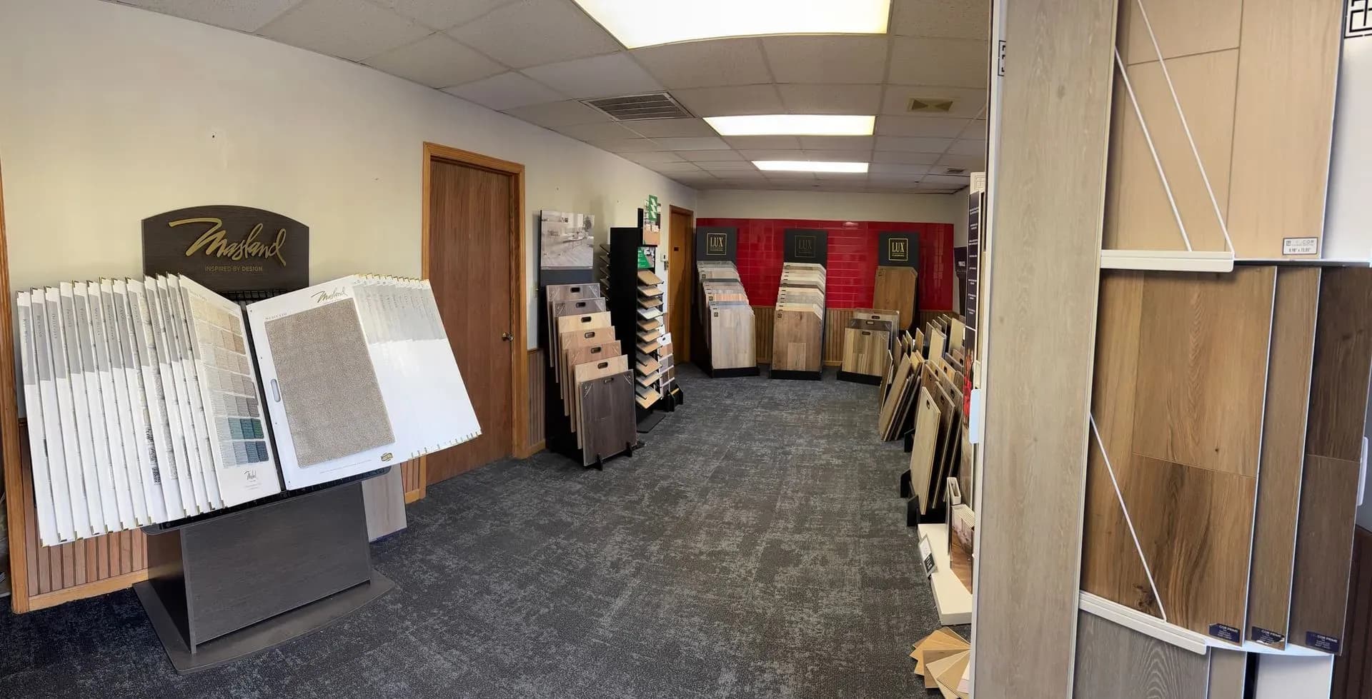 A showroom displaying various flooring samples, including carpet and wood. Samples are neatly arranged on stands along the walls, under bright overhead lighting.